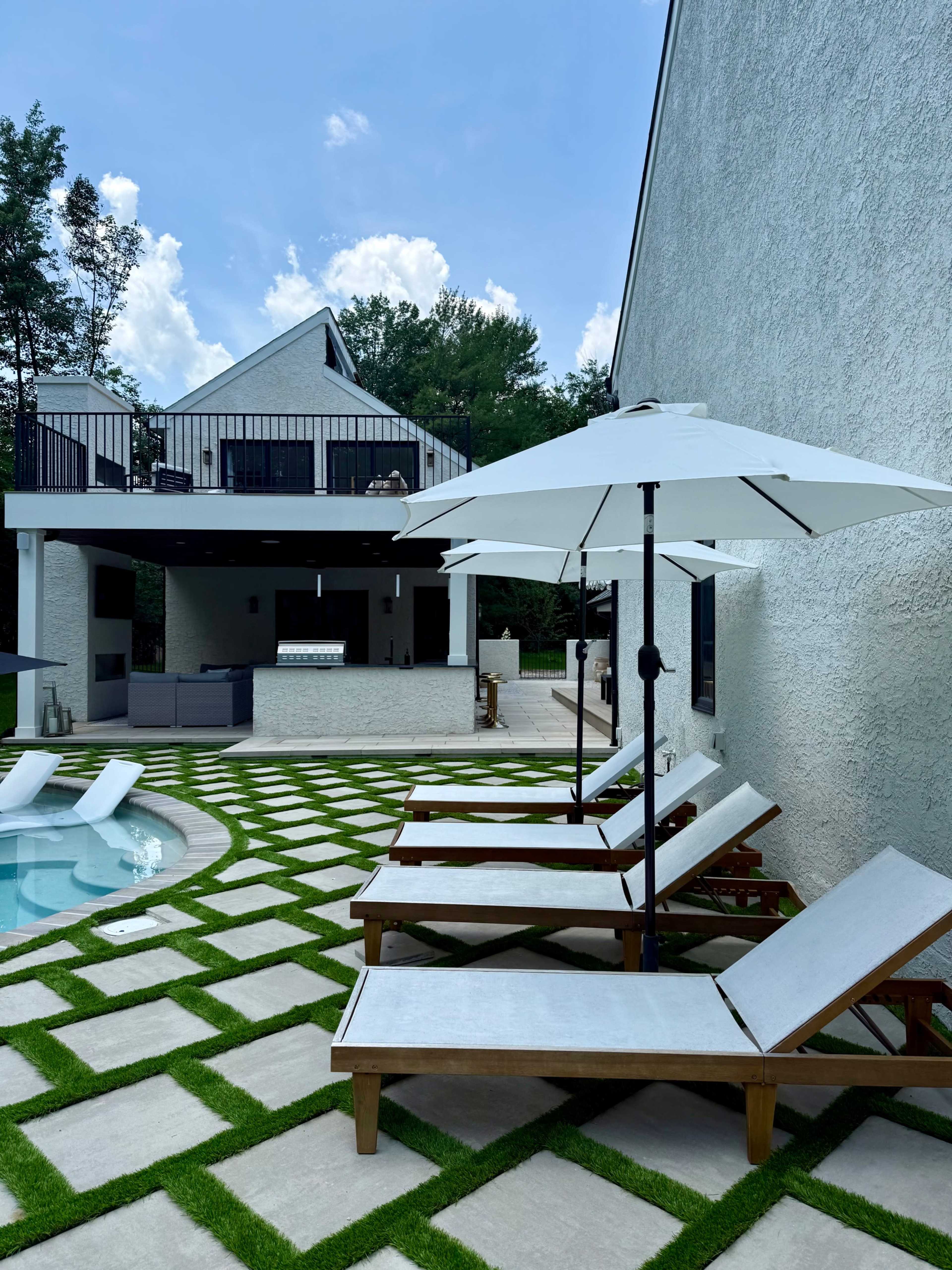 The image shows a modern outdoor space featuring a swimming pool, wooden lounge chairs with white cushions, and an adjacent patio area with an outdoor kitchen.