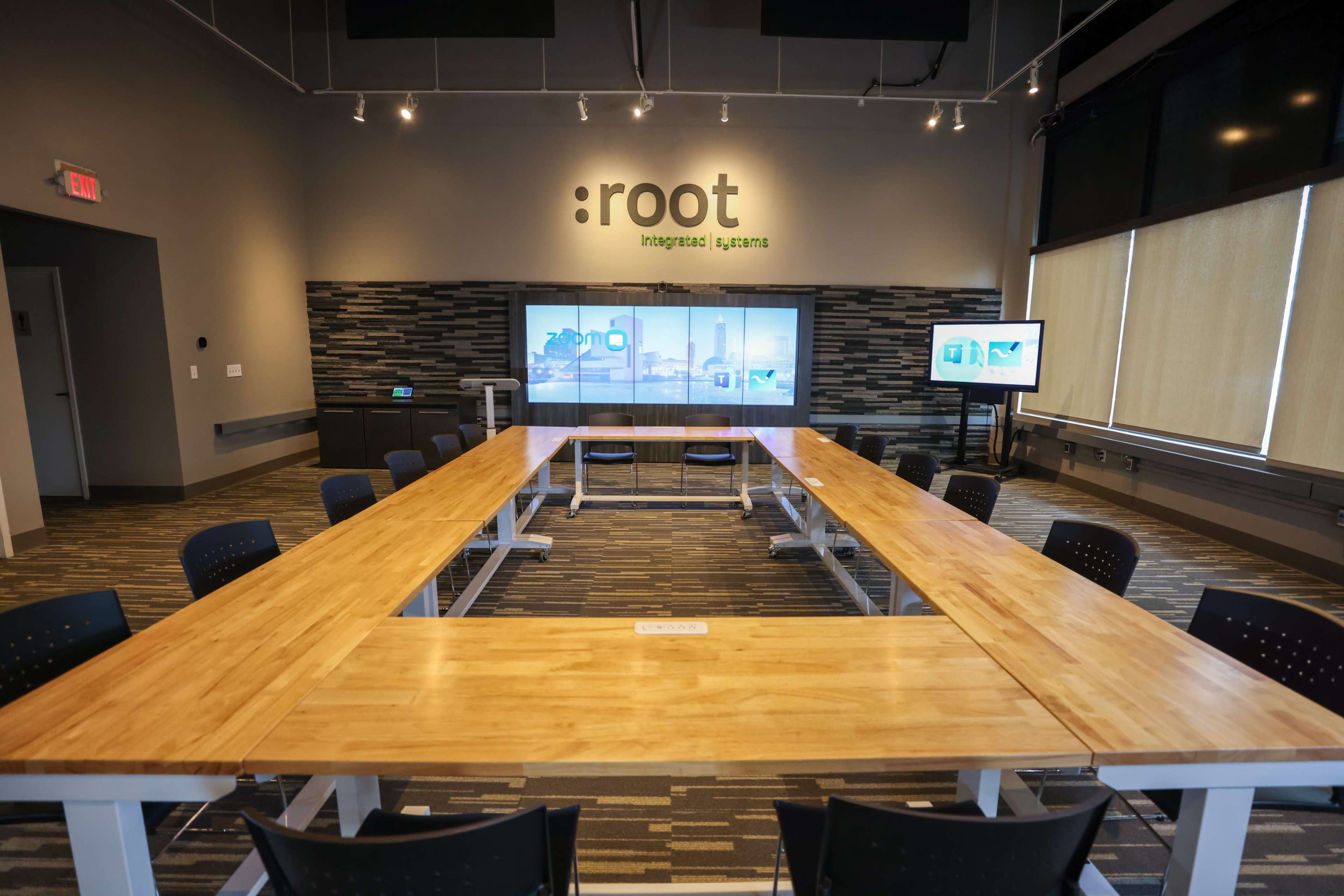 A large conference room features a wooden table surrounded by black chairs, with a screen displaying graphics on one wall.