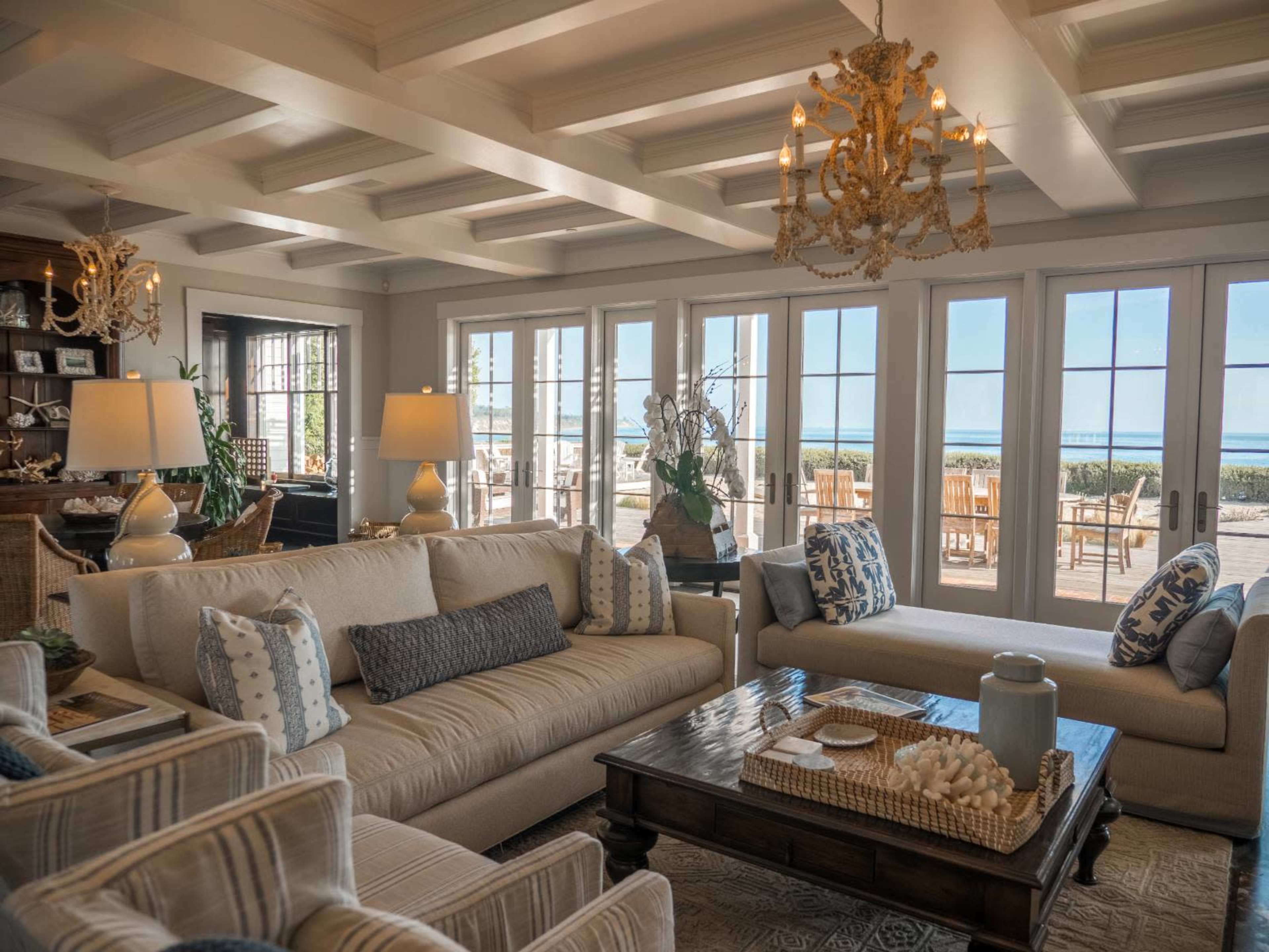 The image shows a bright living room with a large beige sofa, patterned armchairs, and a wooden coffee table, featuring large windows that open to a view of an outdoor dining area and the water.