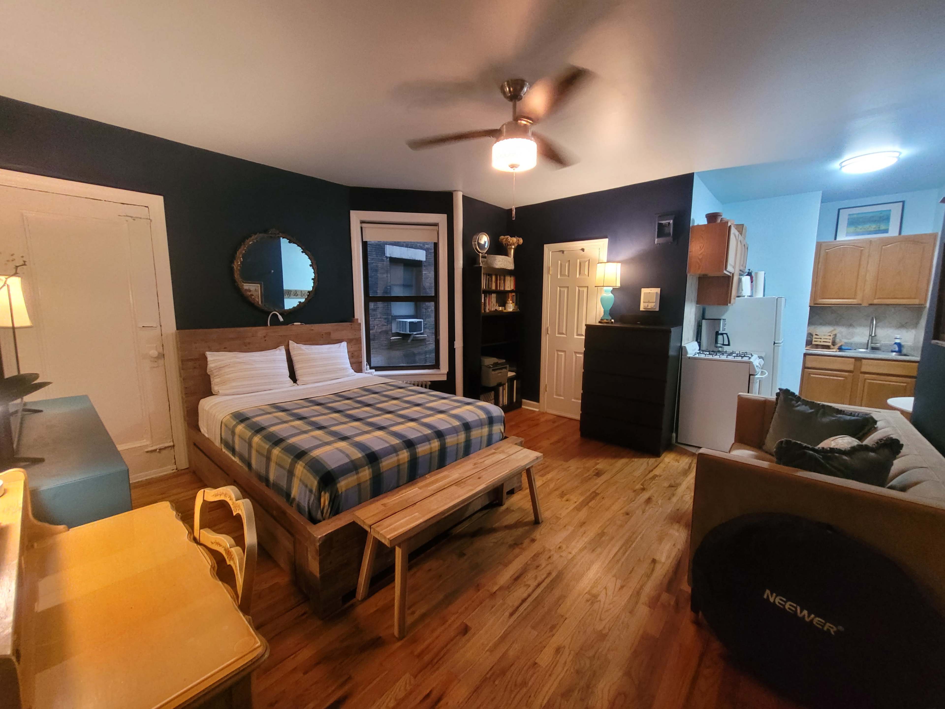 The image shows a small studio apartment featuring a bed with a checkered blanket, a kitchenette, a sofa, a wooden bench, and a ceiling fan.