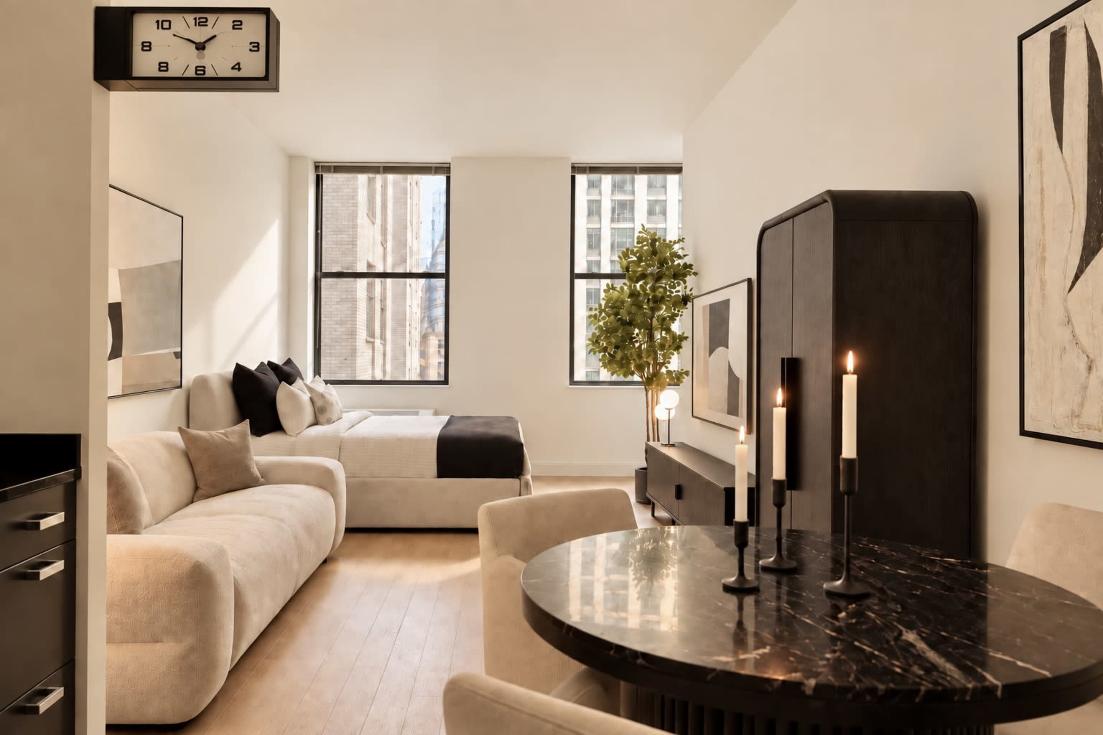 The image shows a modern studio apartment featuring a light-colored sofa, a bed with dark bedding, a round dining table with candles, and large windows allowing natural light.