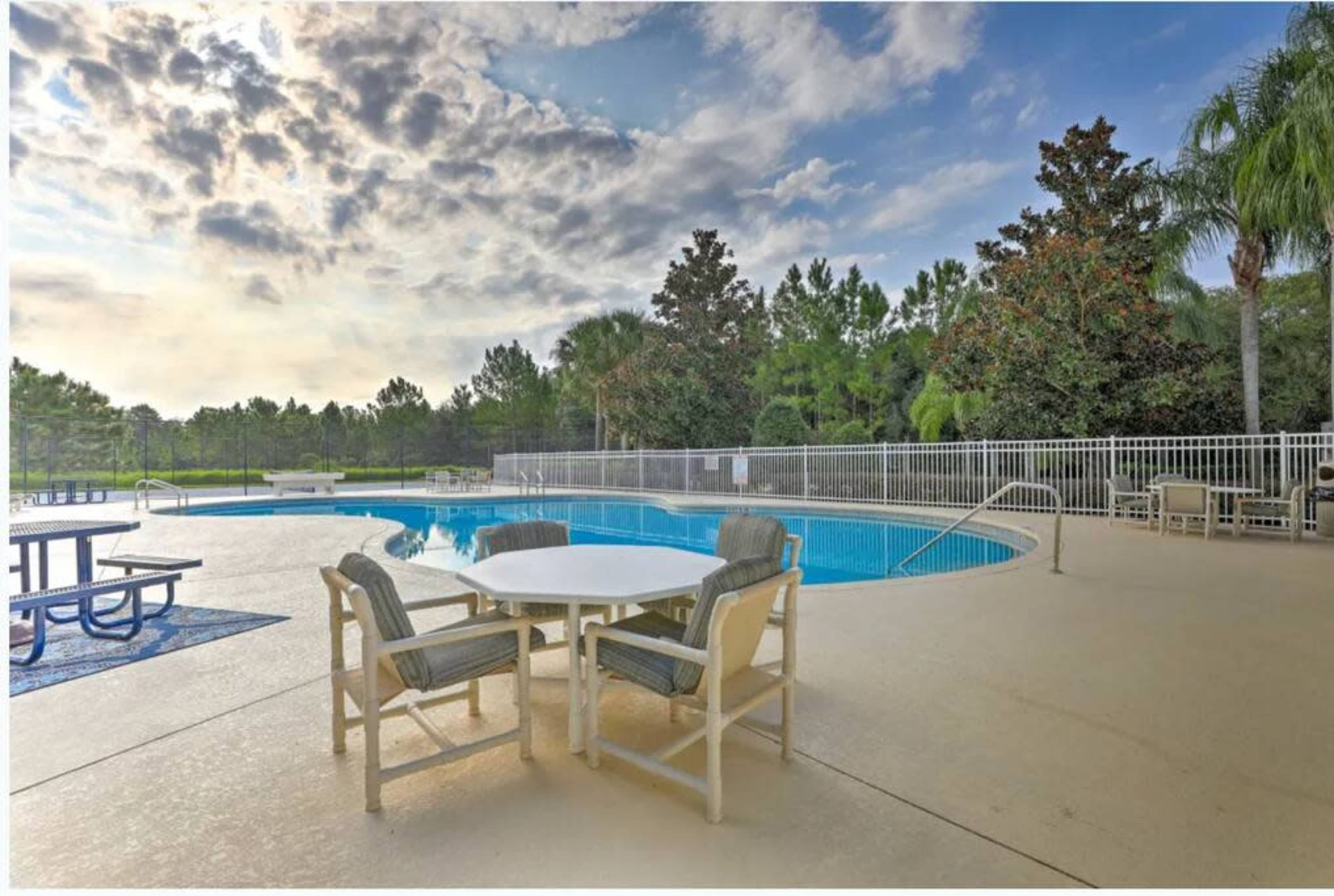 The image shows a swimming pool surrounded by lounge chairs and a dining table in a landscaped outdoor area under a cloudy sky.