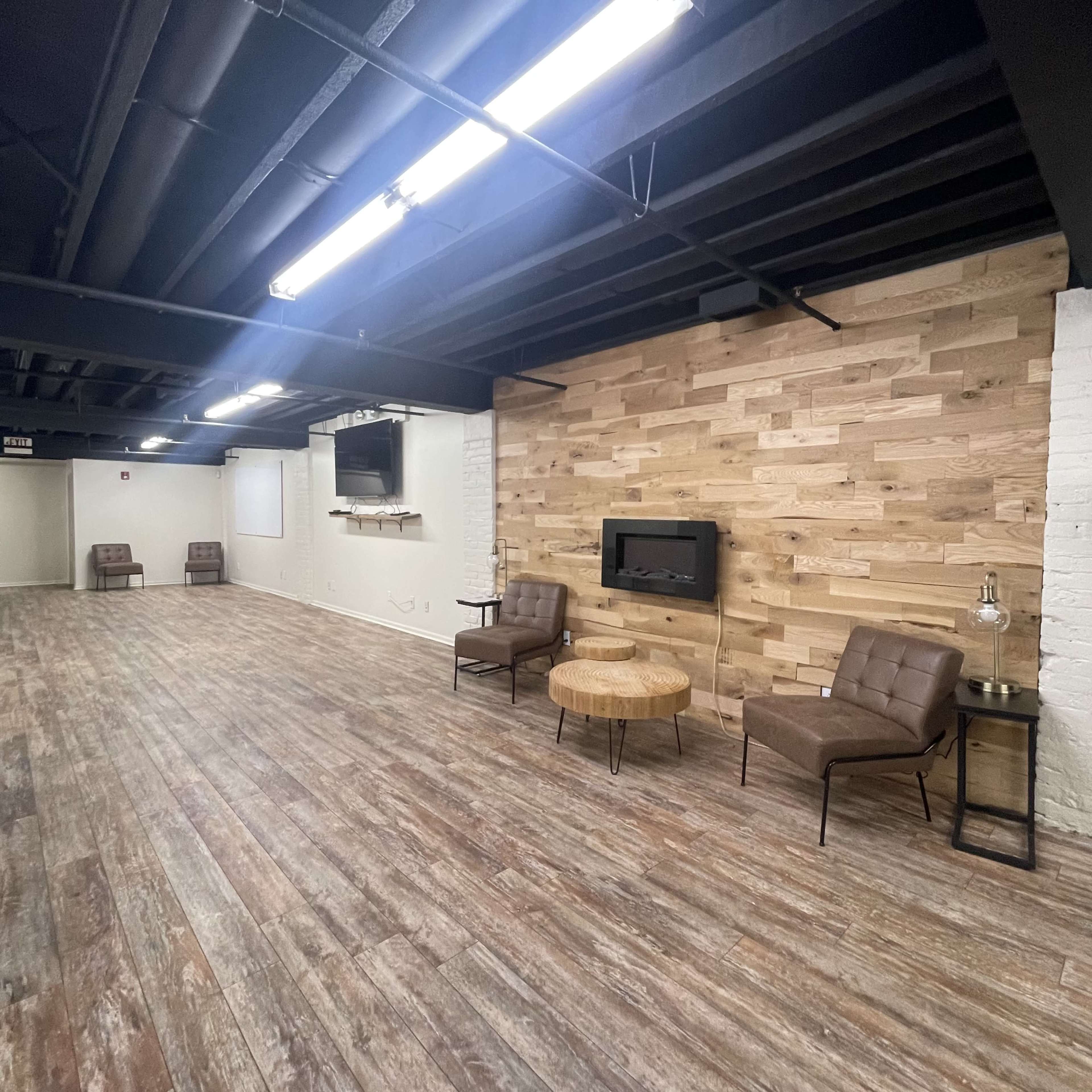 The image shows a spacious room with wooden flooring, featuring a wooden accent wall, two chairs, a coffee table, and mounted wall decor.