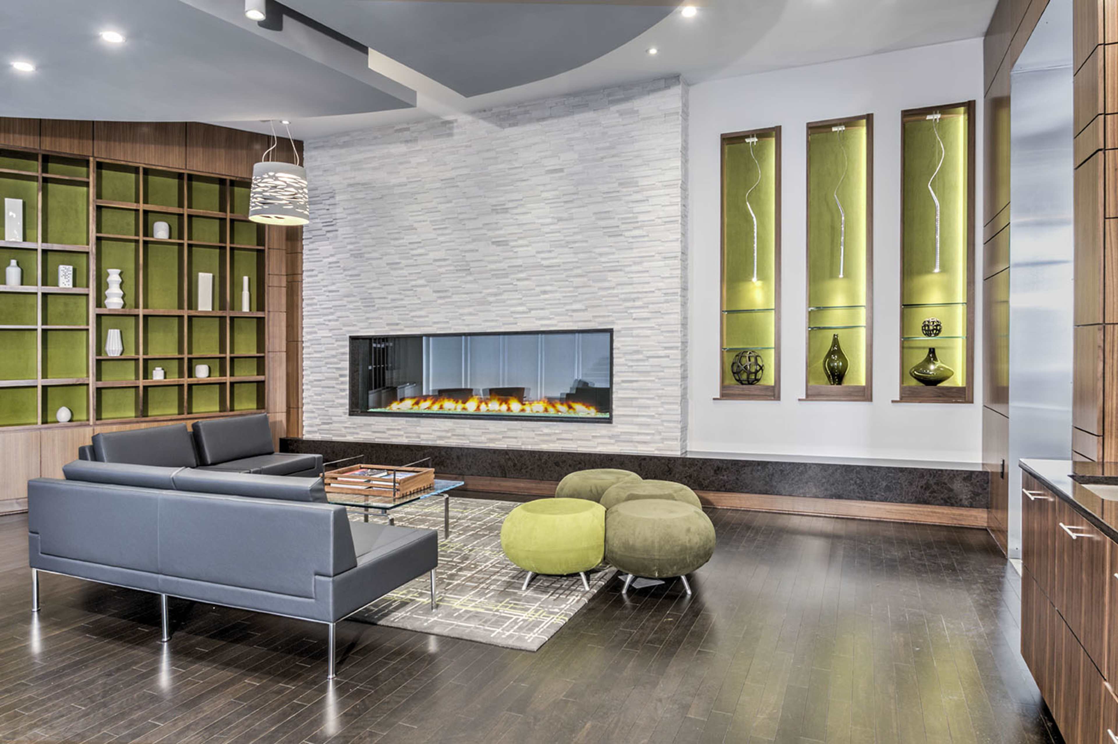 A contemporary living room features a sleek gray sofa, round green poufs, a modern fireplace, and a wall of shelves displaying decorative items.