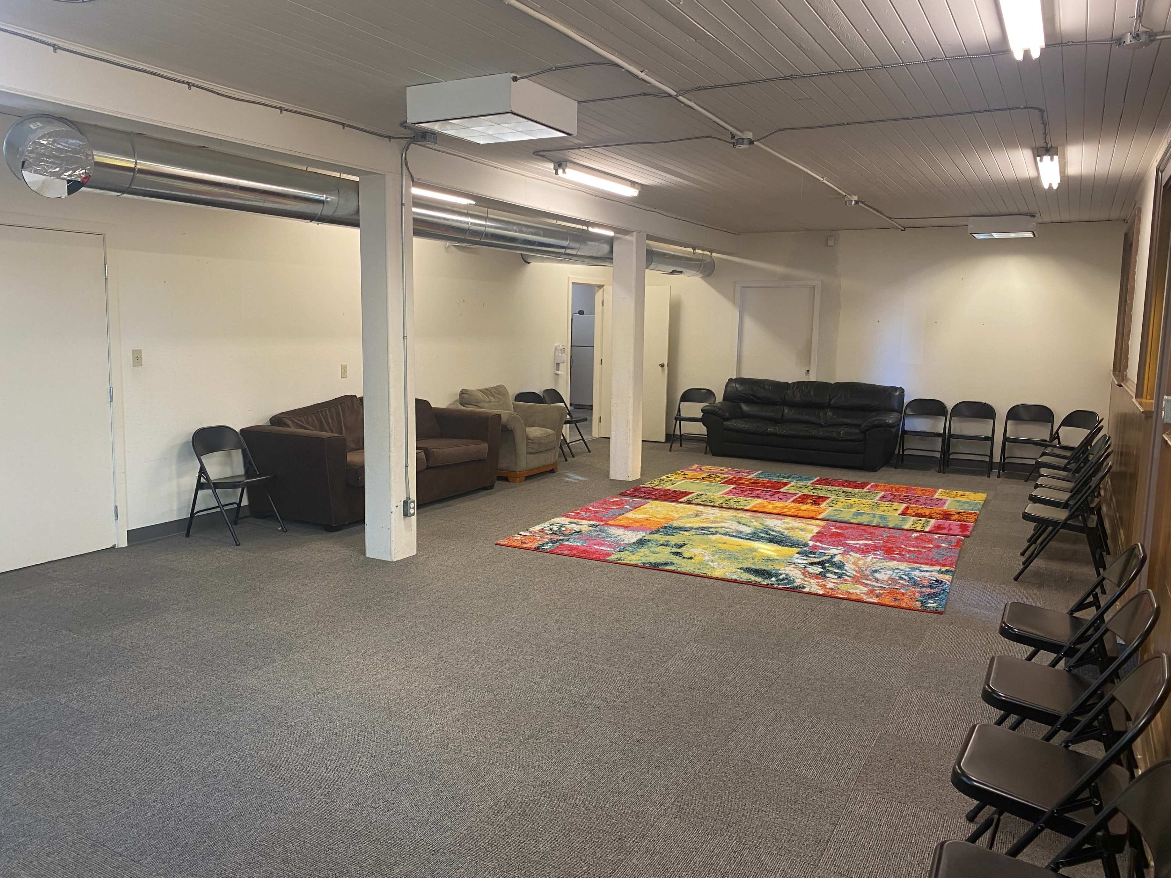 A spacious room with a large colorful rug in the center, surrounded by chairs and a few couches along the walls.