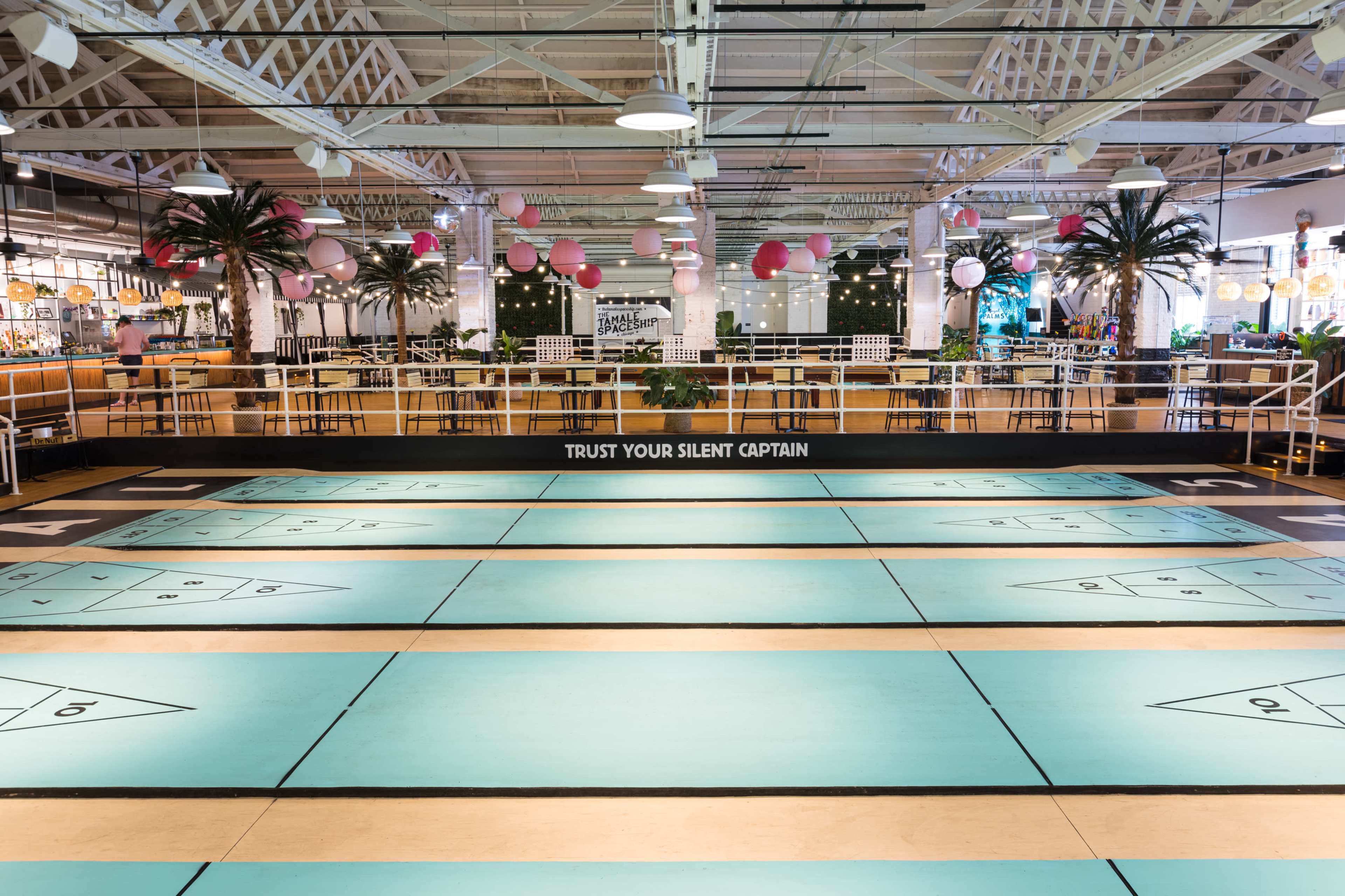 The image shows an indoor entertainment venue featuring a shuffleboard court with a colorful teal and wood floor, surrounded by dining tables and decorations.