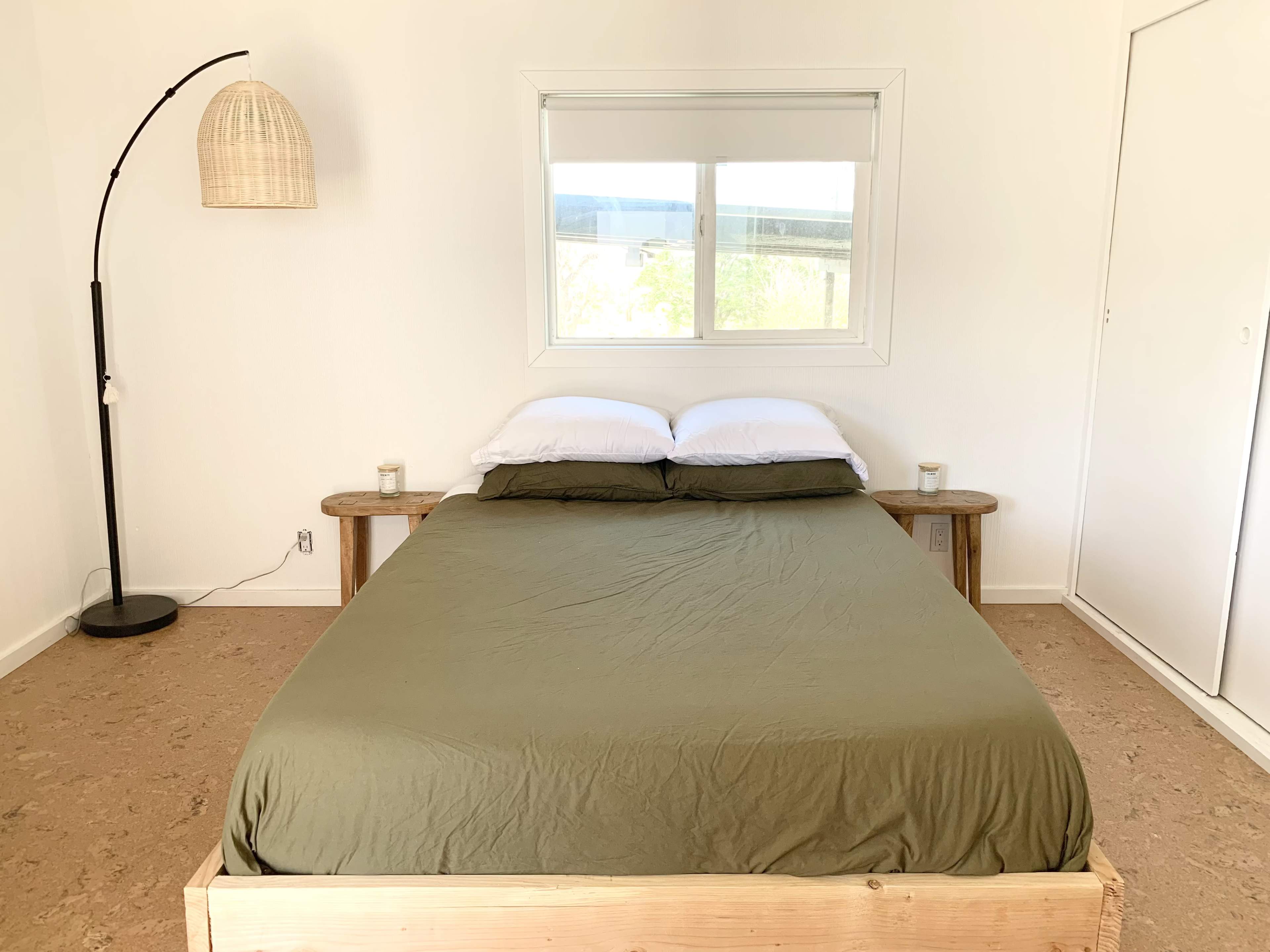 A simple bedroom features a wooden bed with an olive green blanket, two white pillows, and a modern floor lamp, positioned next to a window.