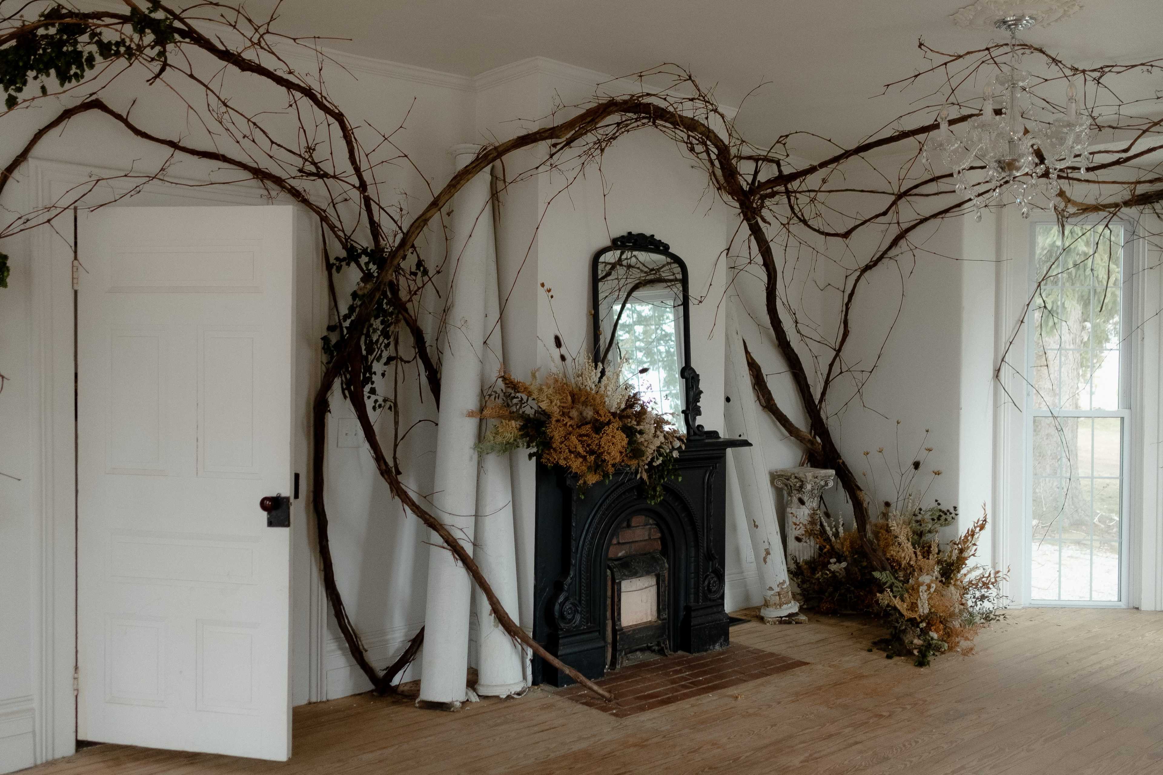 The room features a fireplace surrounded by large, twisted vines and dried floral arrangements, with a mirror reflecting the space.
