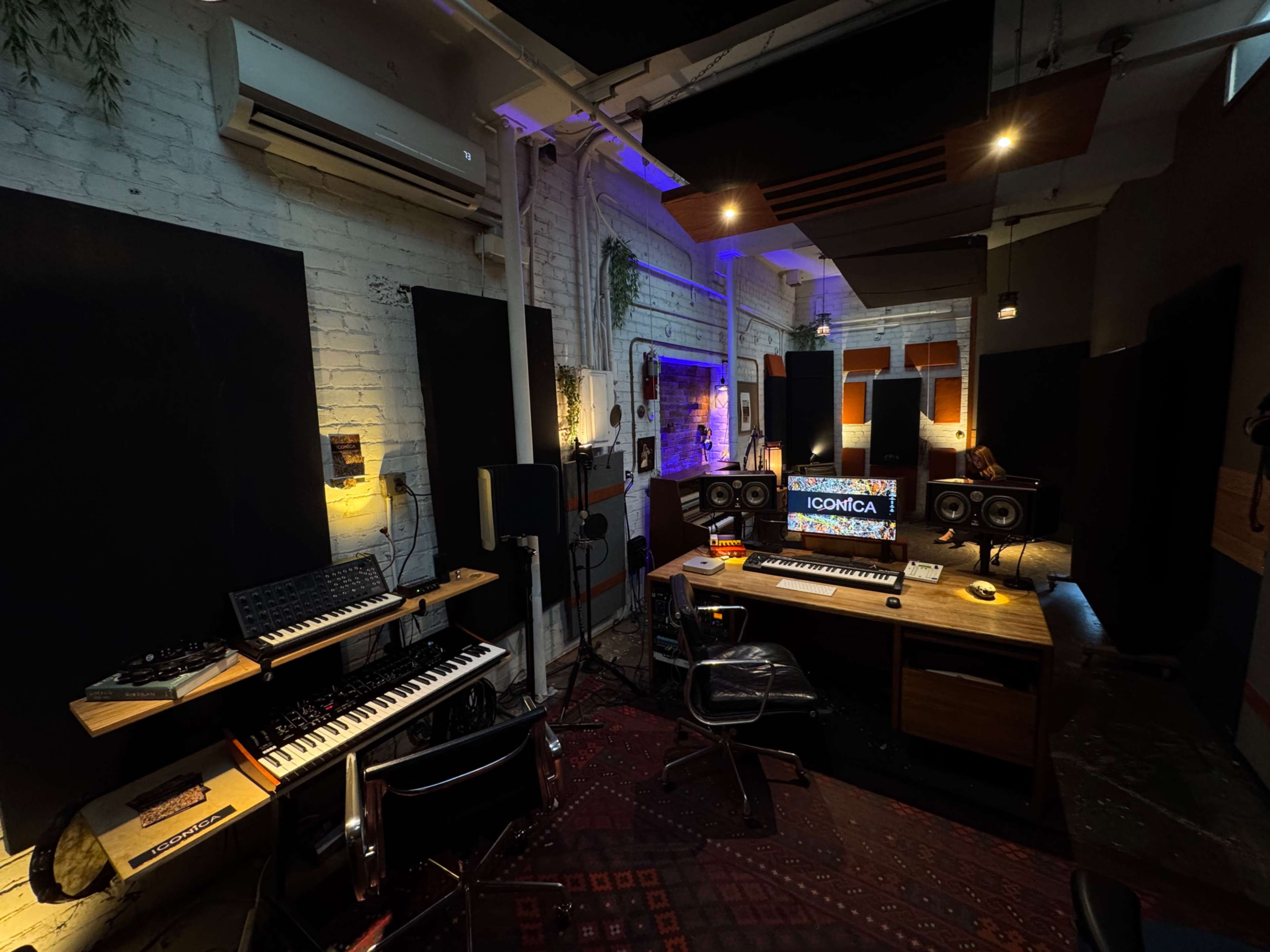 The image shows a modern music studio featuring a computer workstation, keyboards, studio monitors, and soundproof wall panels.