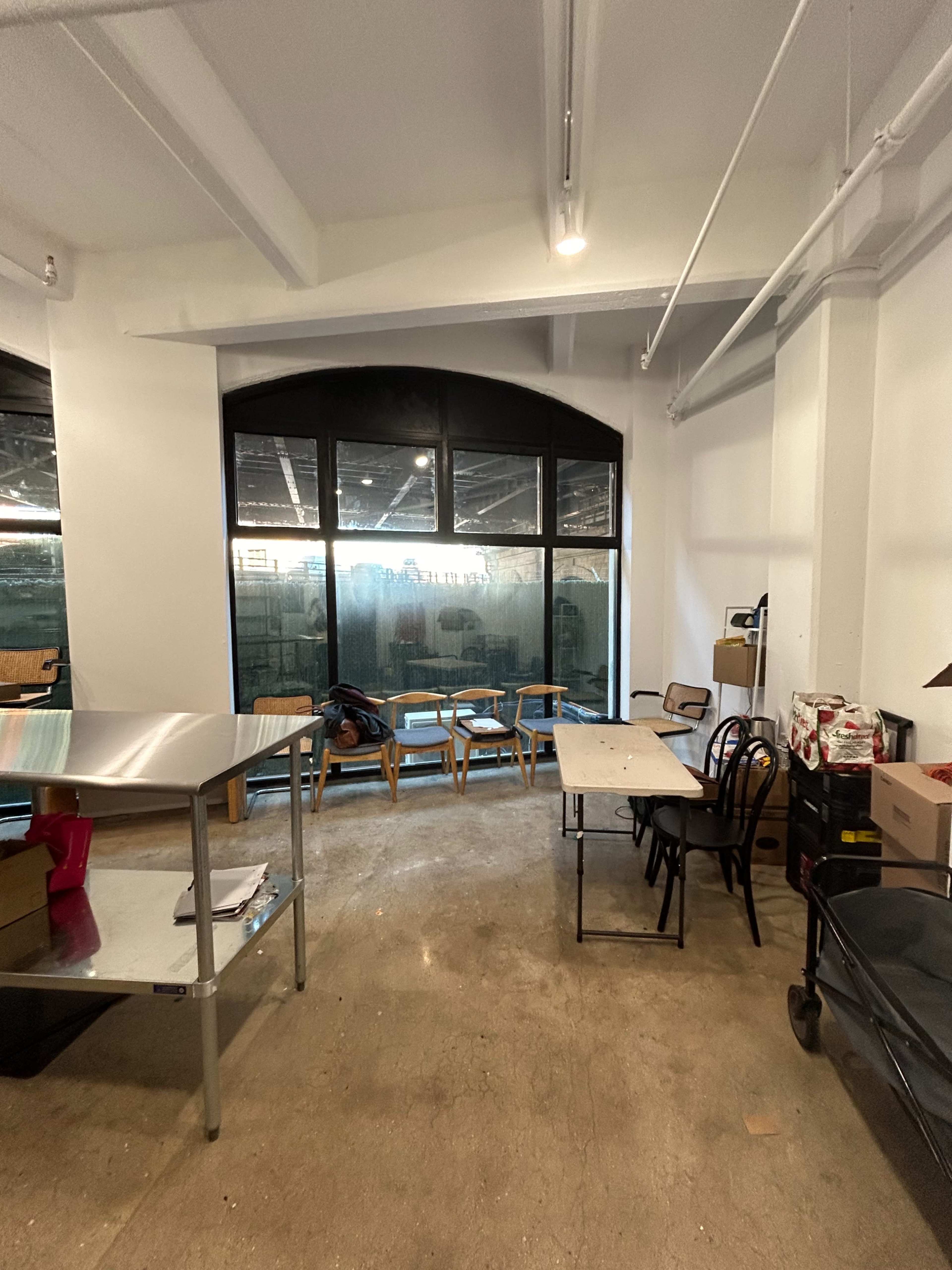 The image shows an empty room with tables and chairs arranged near a large window that overlooks an outdoor area.