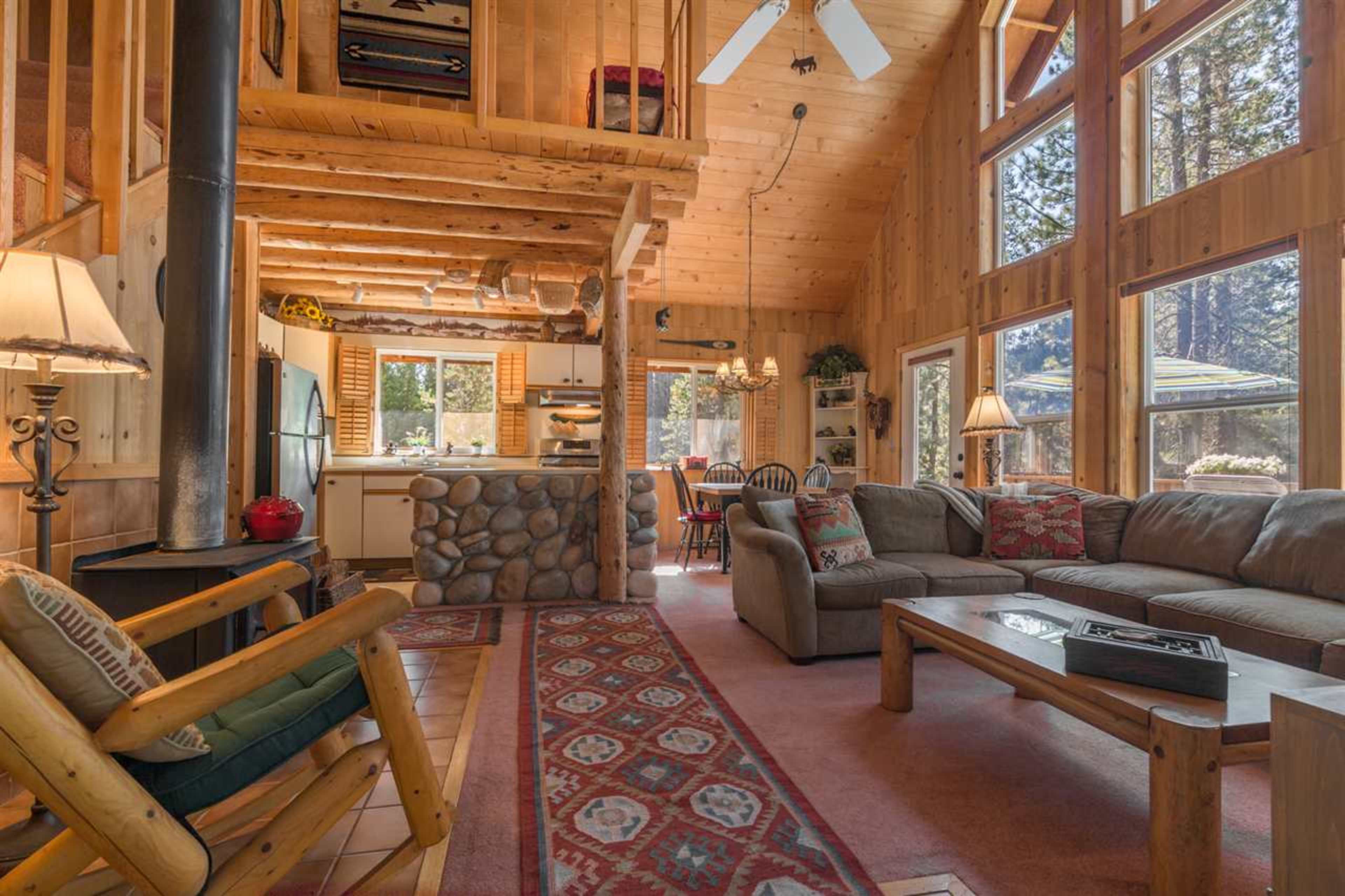 The image shows a cozy, wooden cabin living room with large windows, a stone fireplace, and a kitchen area visible in the background.