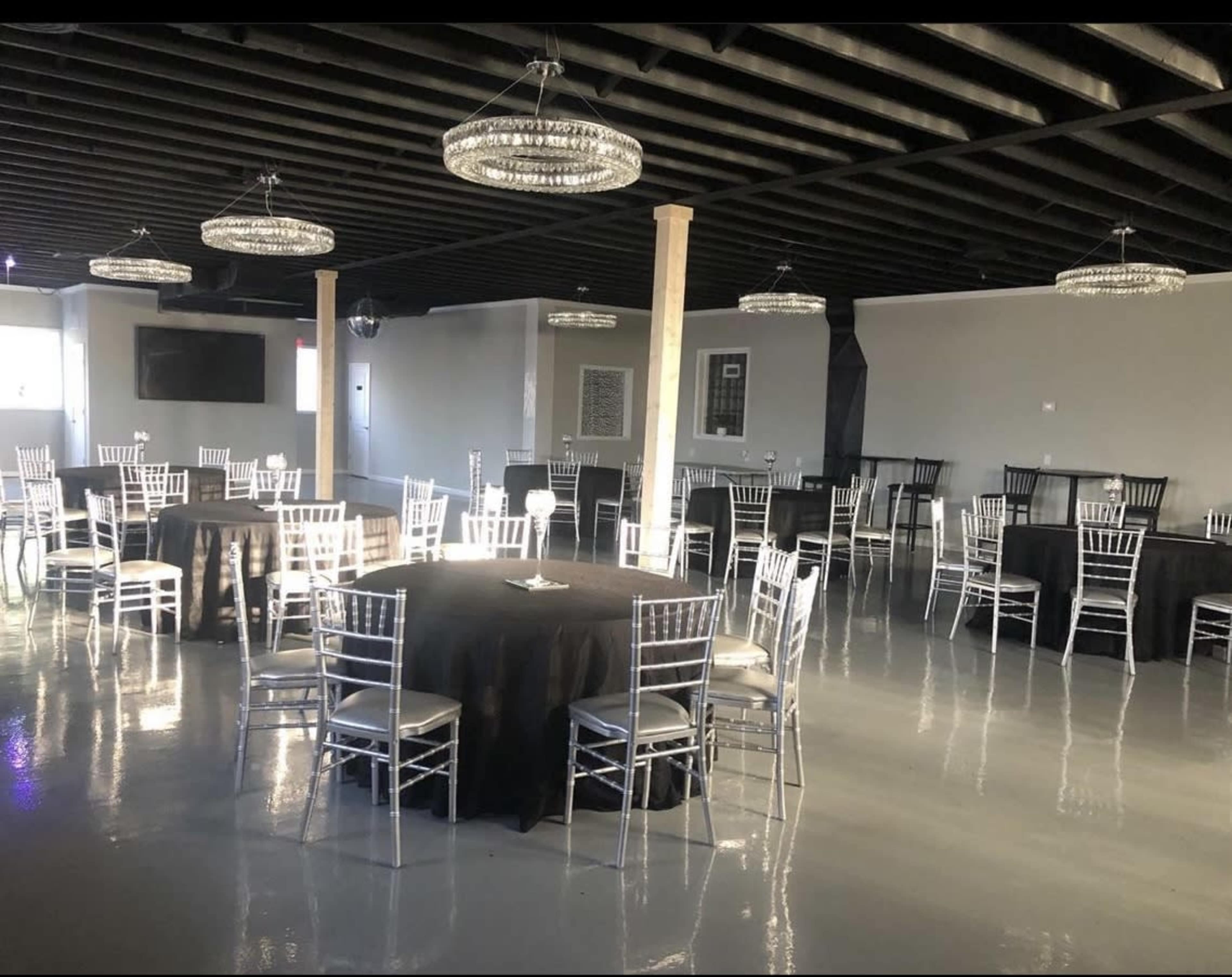 A spacious event venue with black round tables surrounded by silver chiavari chairs and modern chandelier lighting.