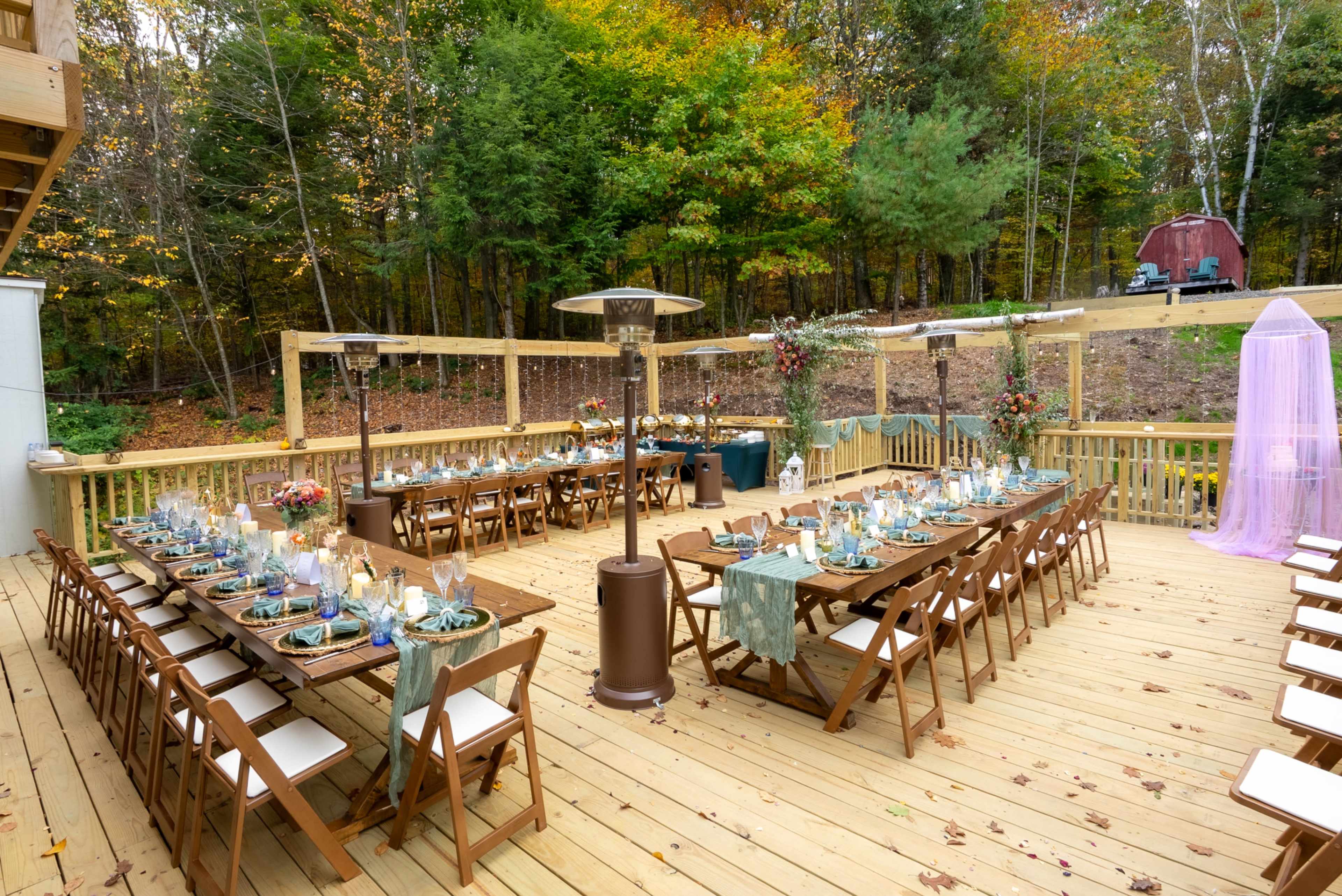 The image shows a beautifully arranged outdoor dining space with tables set for a gathering, surrounded by trees in a scenic fall setting.