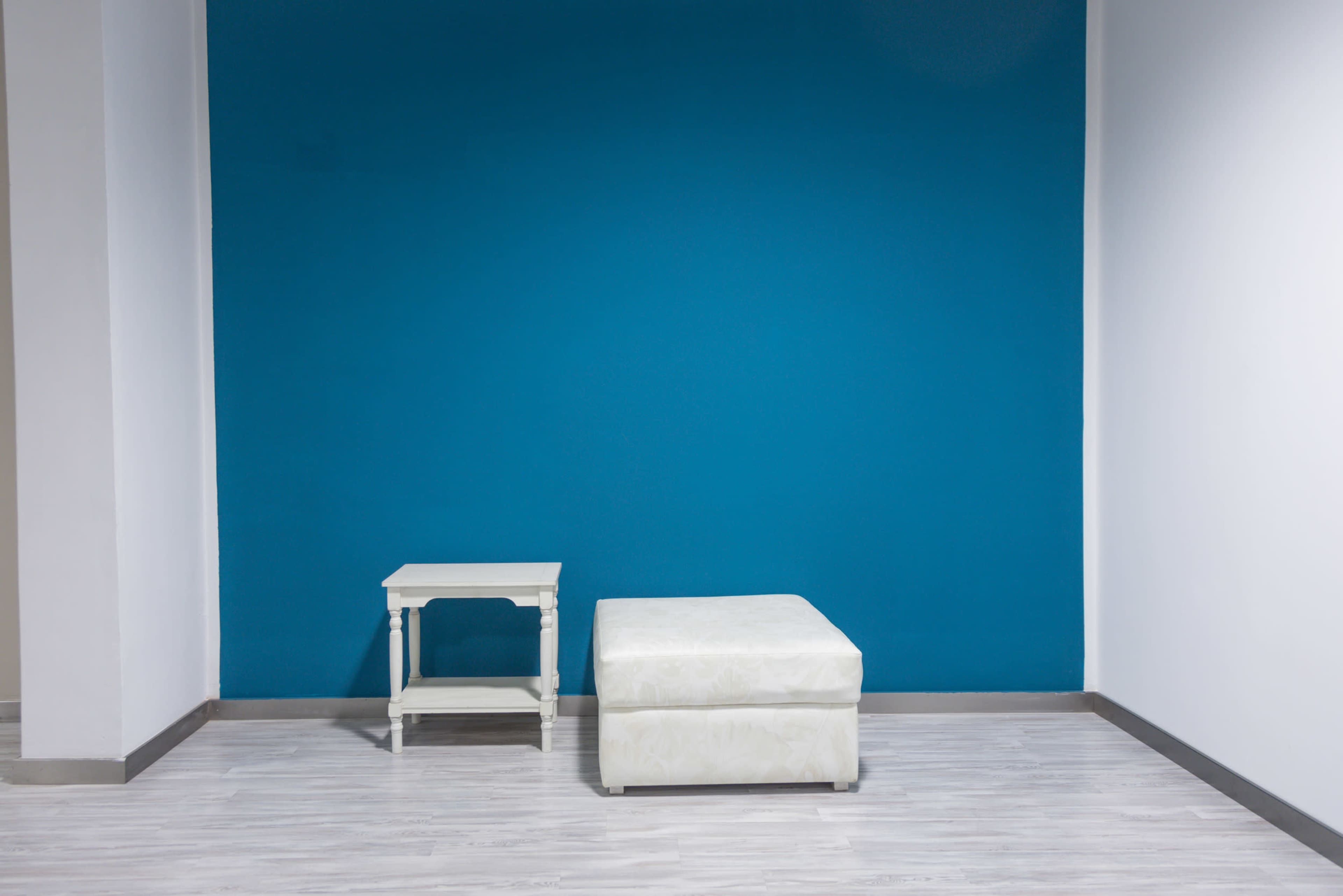 A minimalist interior with a blue wall, a small white side table, and a white ottoman on a light wood floor.