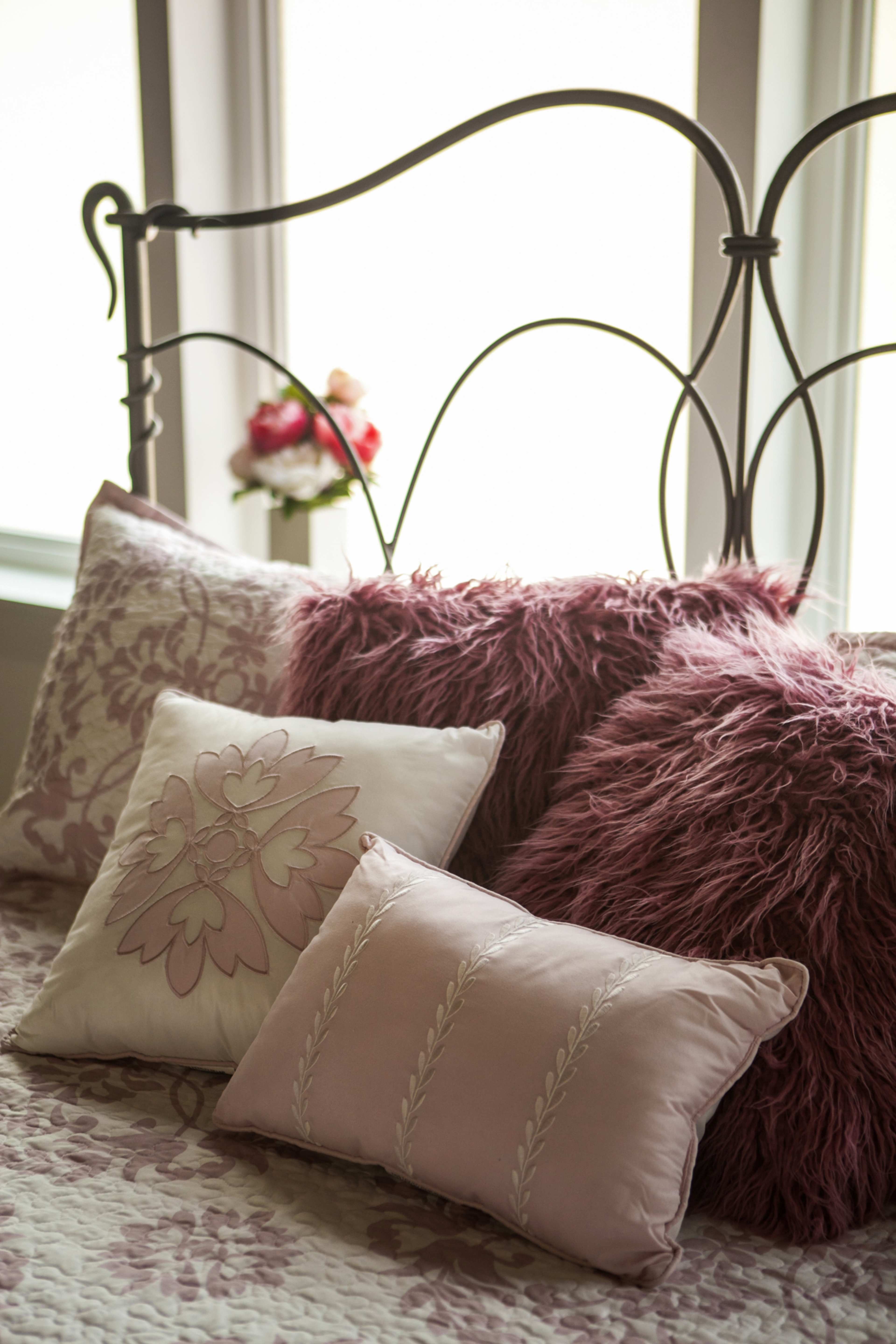 A bed with decorative pillows arranged on a quilted coverlet is positioned near a window with flowers in the background.