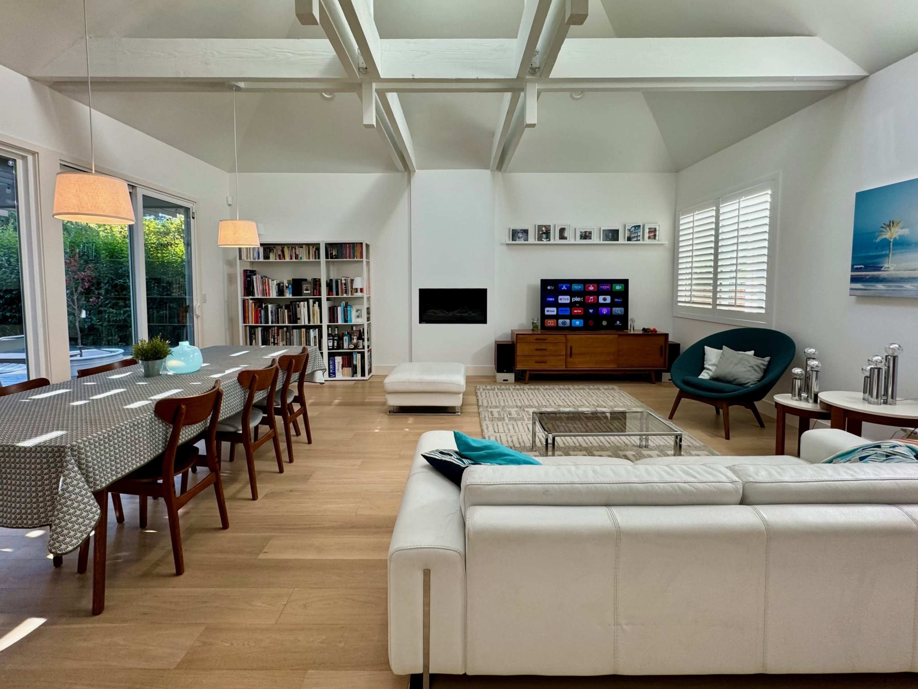 A bright living space features a dining area, a white sofa, a television set, and bookshelves against a backdrop of large windows.