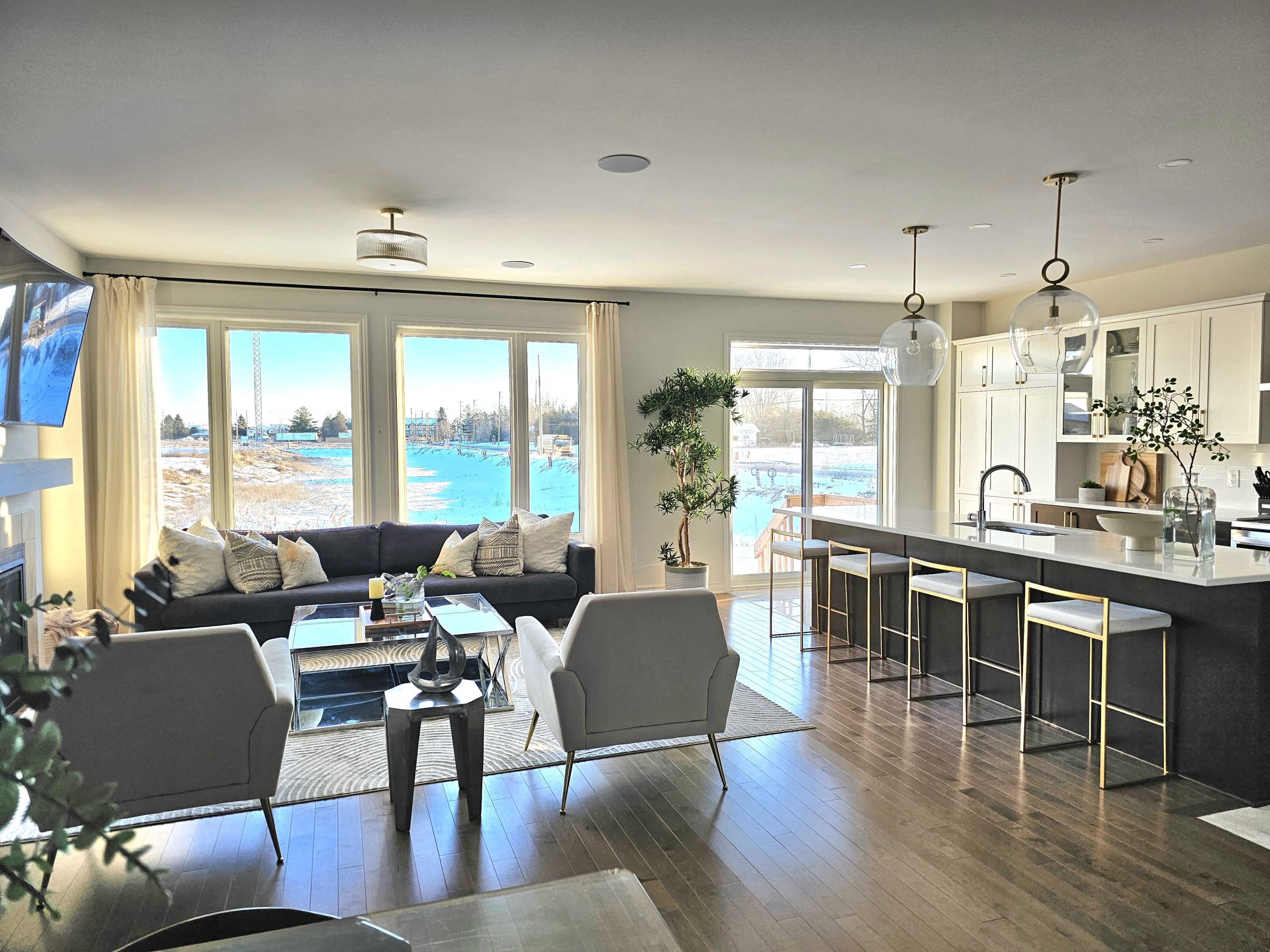 A modern living room features a sectional sofa, two armchairs, and a kitchen island with bar stools, all illuminated by natural light from large windows overlooking a snowy landscape.