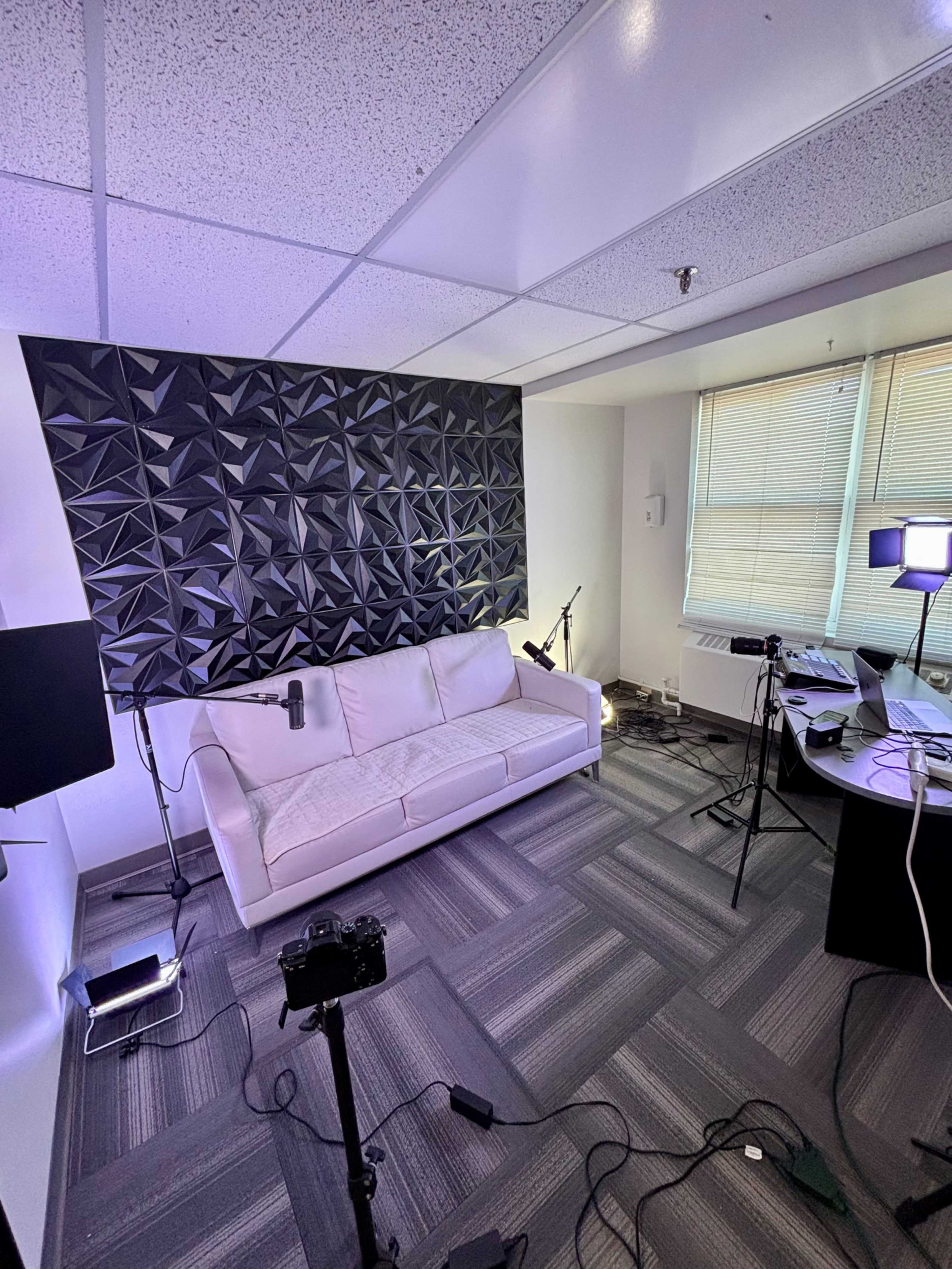 The image shows a modern office space featuring a white couch, a textured black wall, and various camera equipment arranged around a desk with multiple light sources.