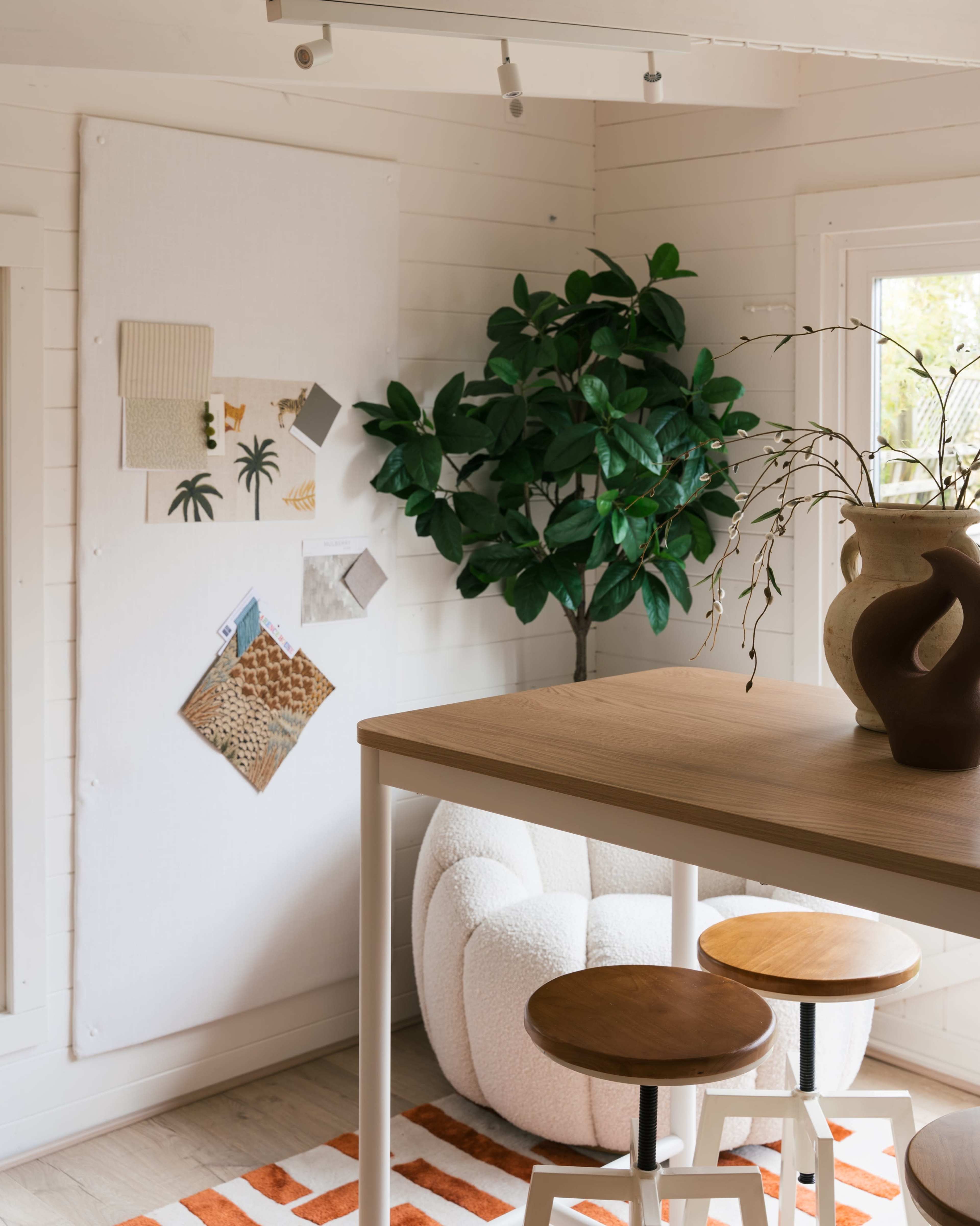 A brightly lit interior featuring a wooden table with two stools, a bulletin board with various pinned notes and images, a decorative plant, and a textured vase on the table.
