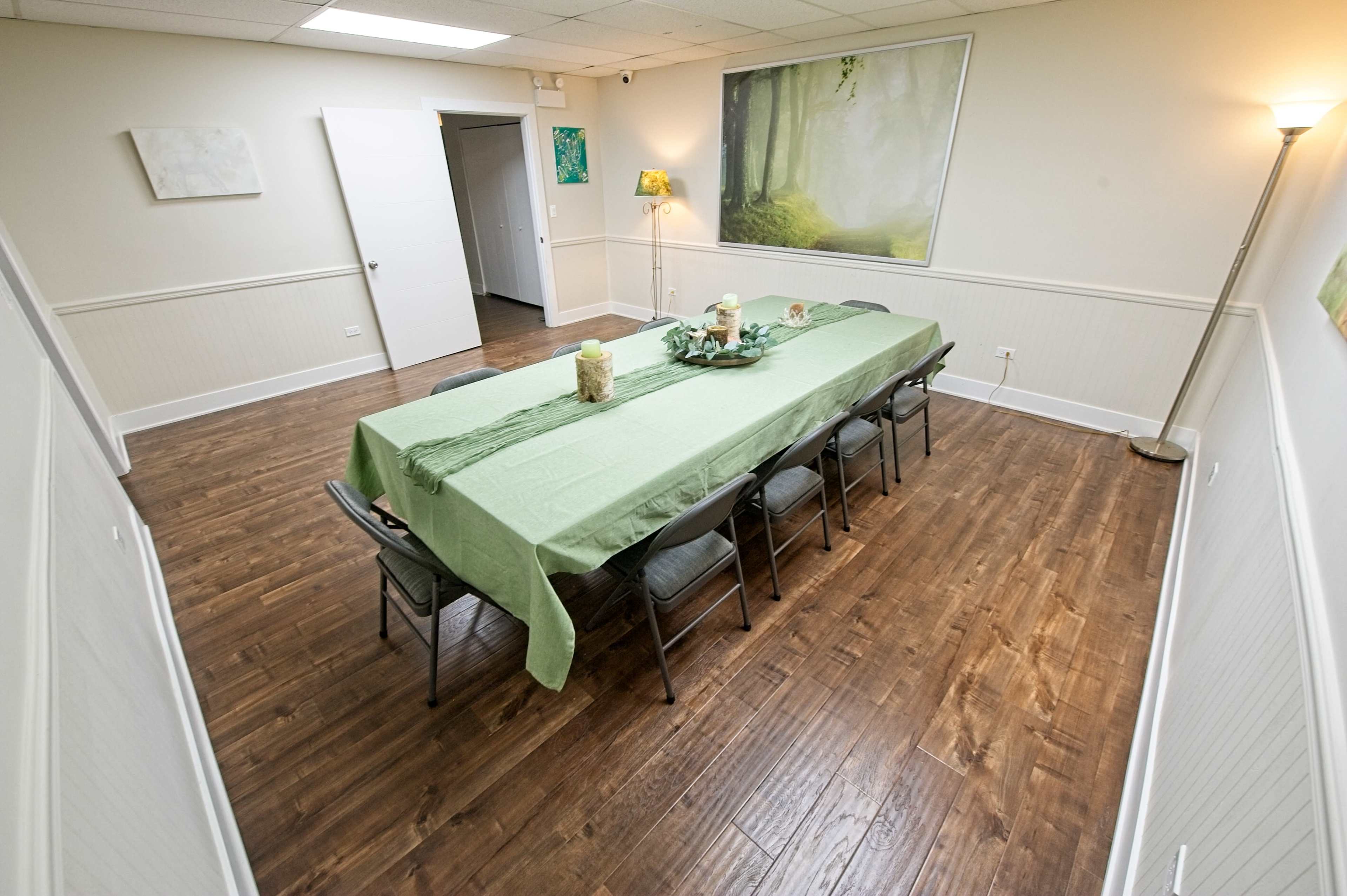 The image shows a spacious meeting room with a long green table set for a gathering, surrounded by folding chairs, and decorated with a floral centerpiece.