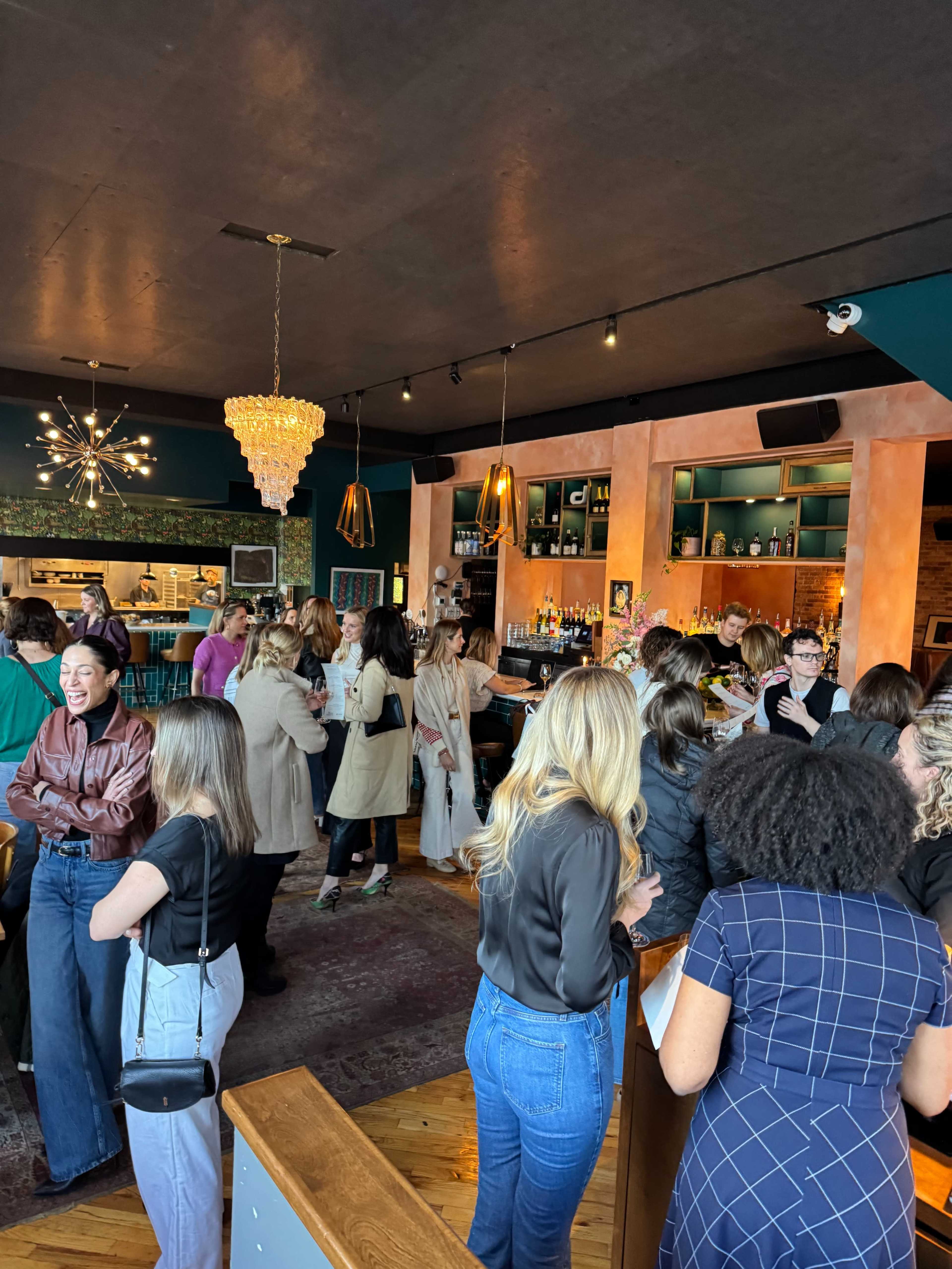 A vibrant gathering of people is taking place in a stylish bar, featuring a mix of conversations and a well-stocked bar in the background.