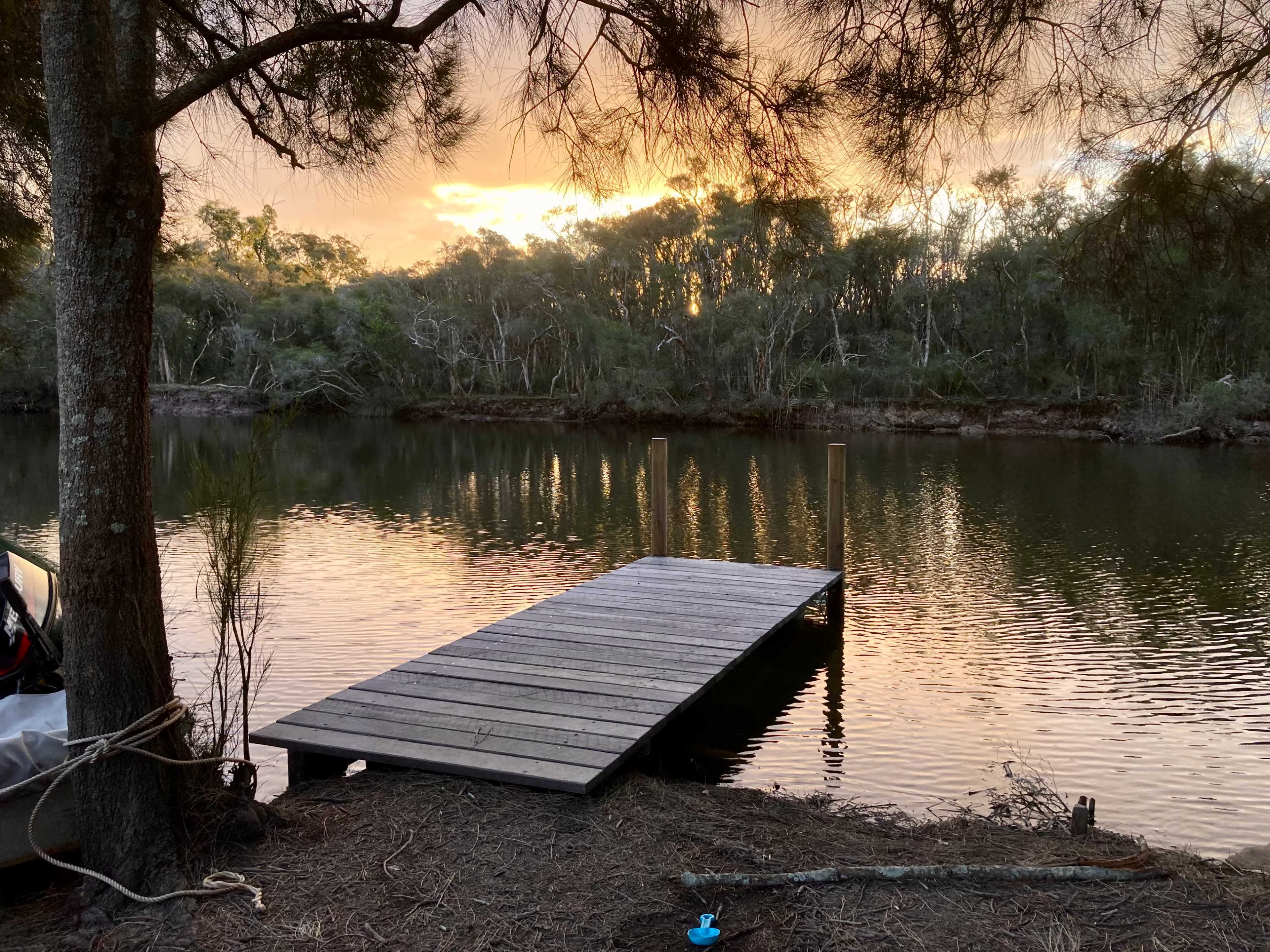 A wooden dock extends into a calm river, with trees lining the shore and a sunset reflecting on the water's surface.