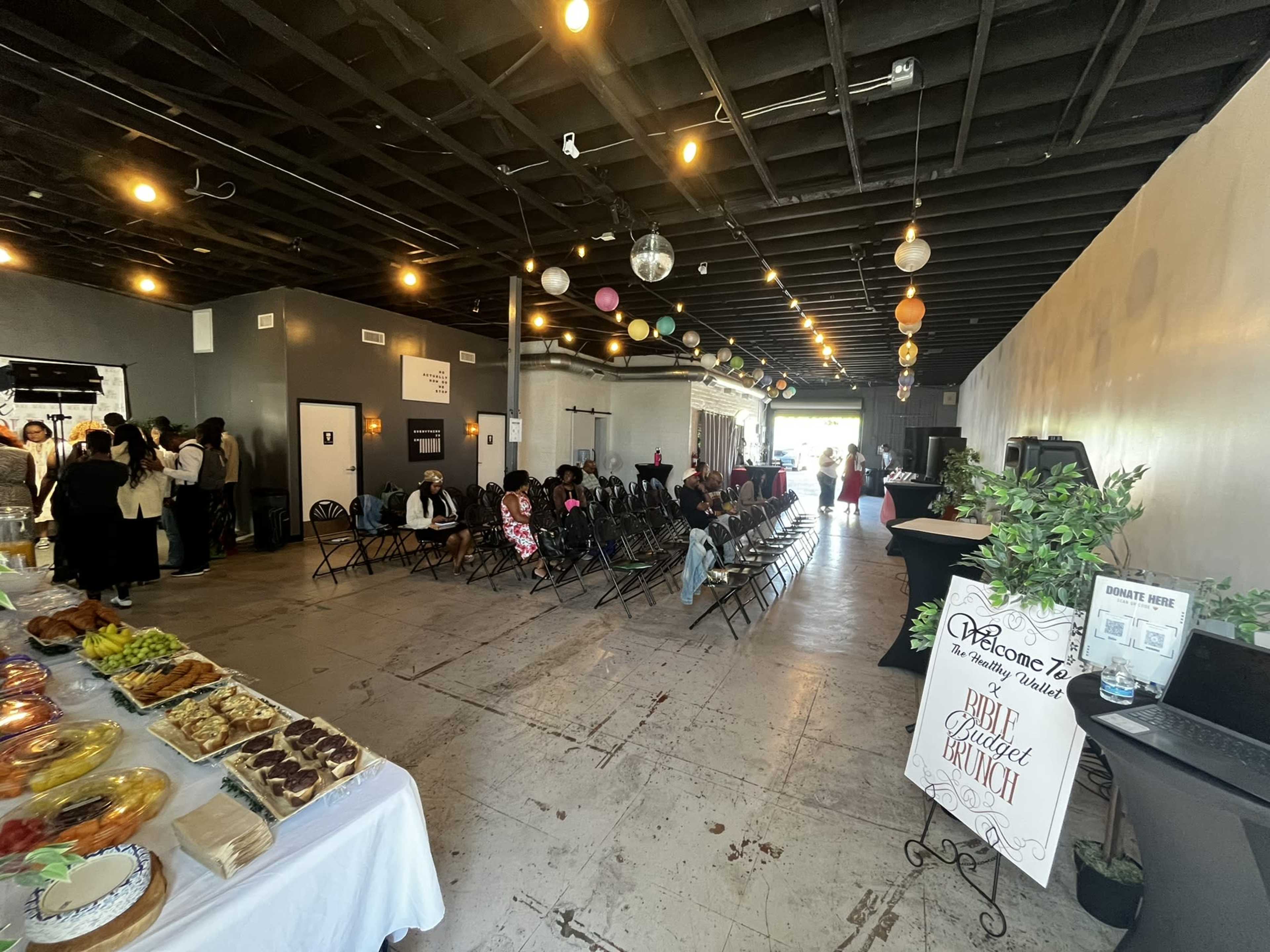 The image shows a spacious event venue with a seating area filled with chairs and tables displaying food, surrounded by attendees engaged in conversation.