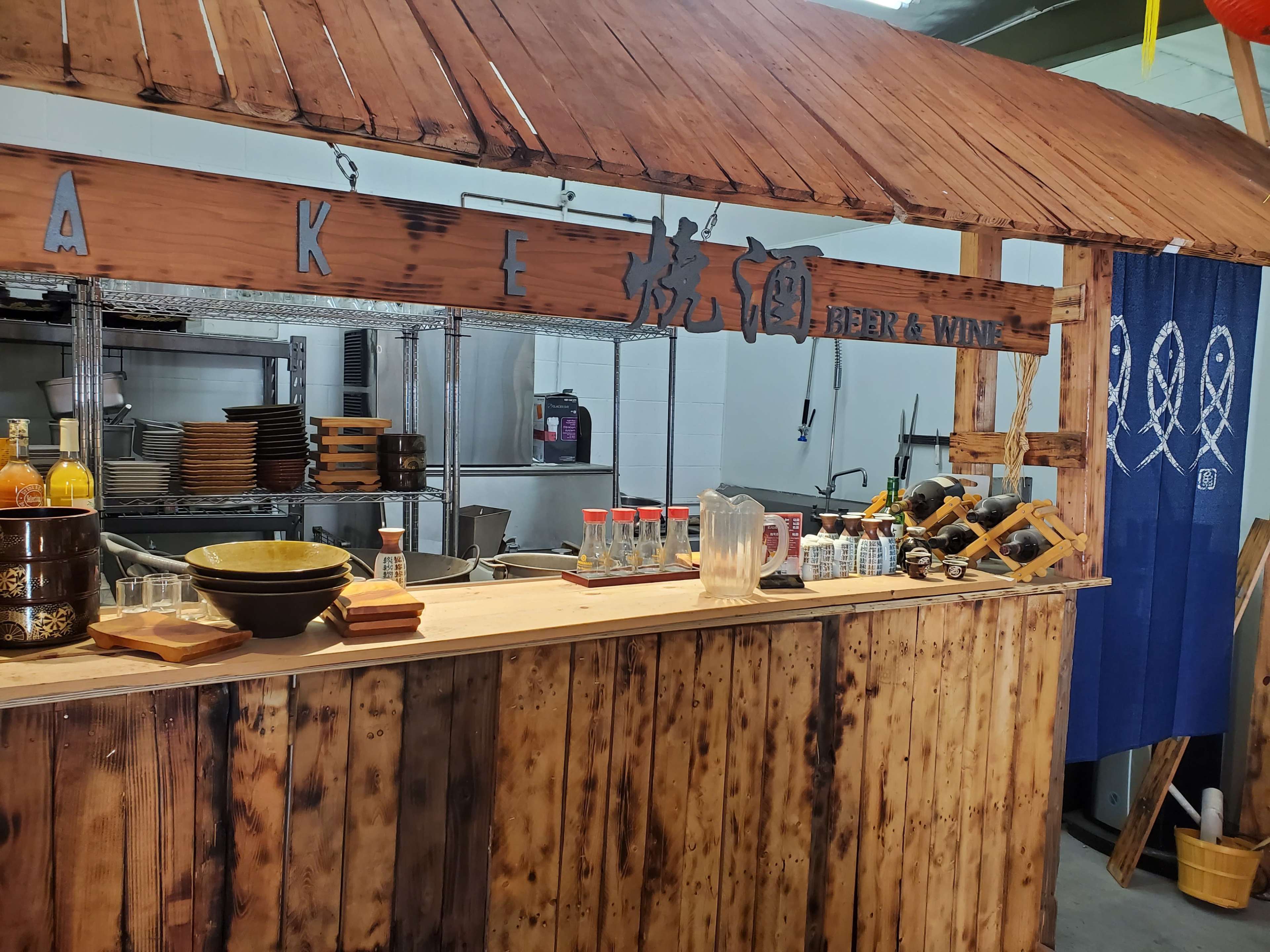 A rustic wooden bar setup with signage for "Beer & Wine," surrounded by kitchen equipment and shelves in an industrial space.