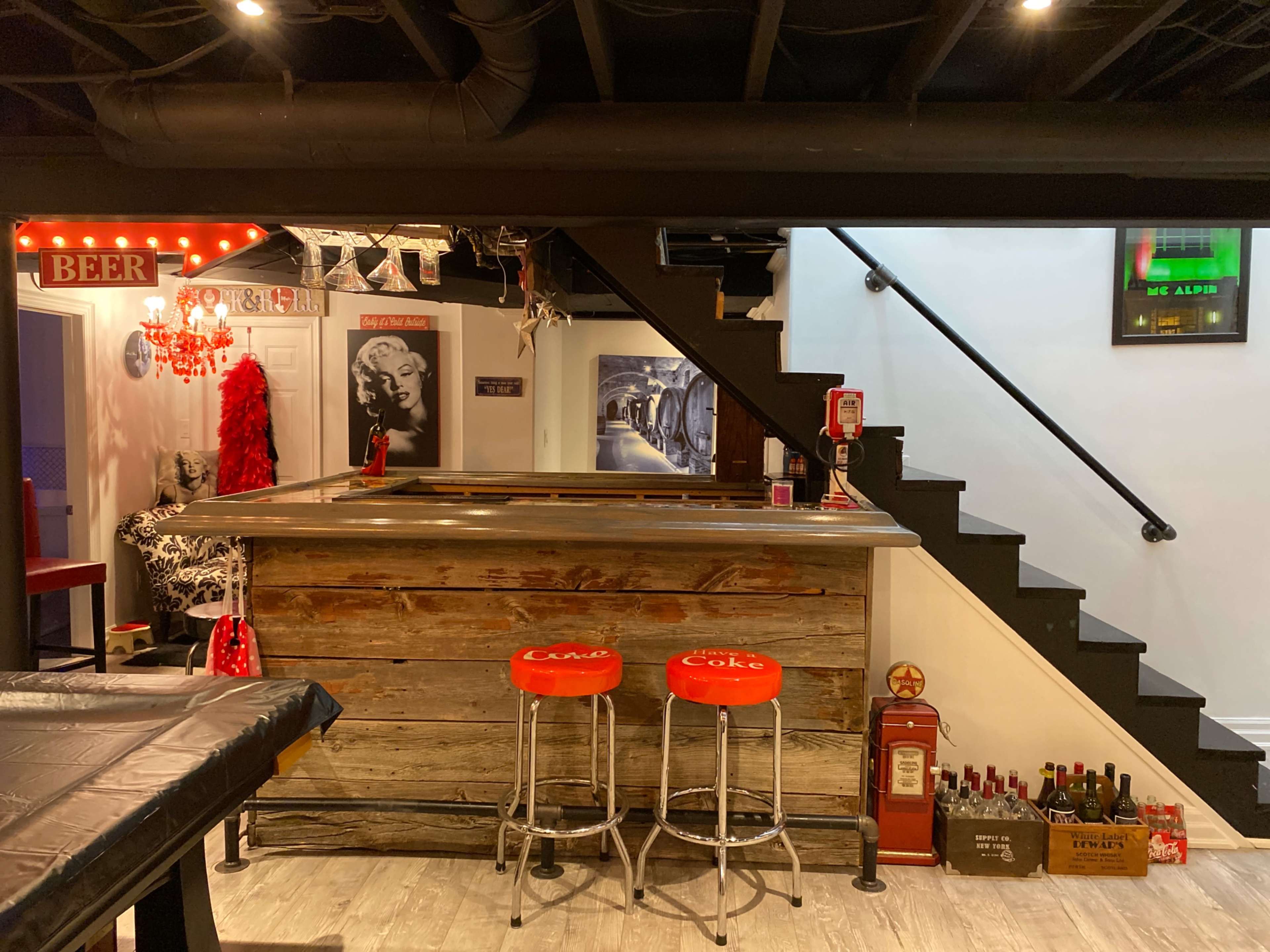 A basement bar area with a wooden bar counter, red bar stools, and a staircase leading to an upper level.