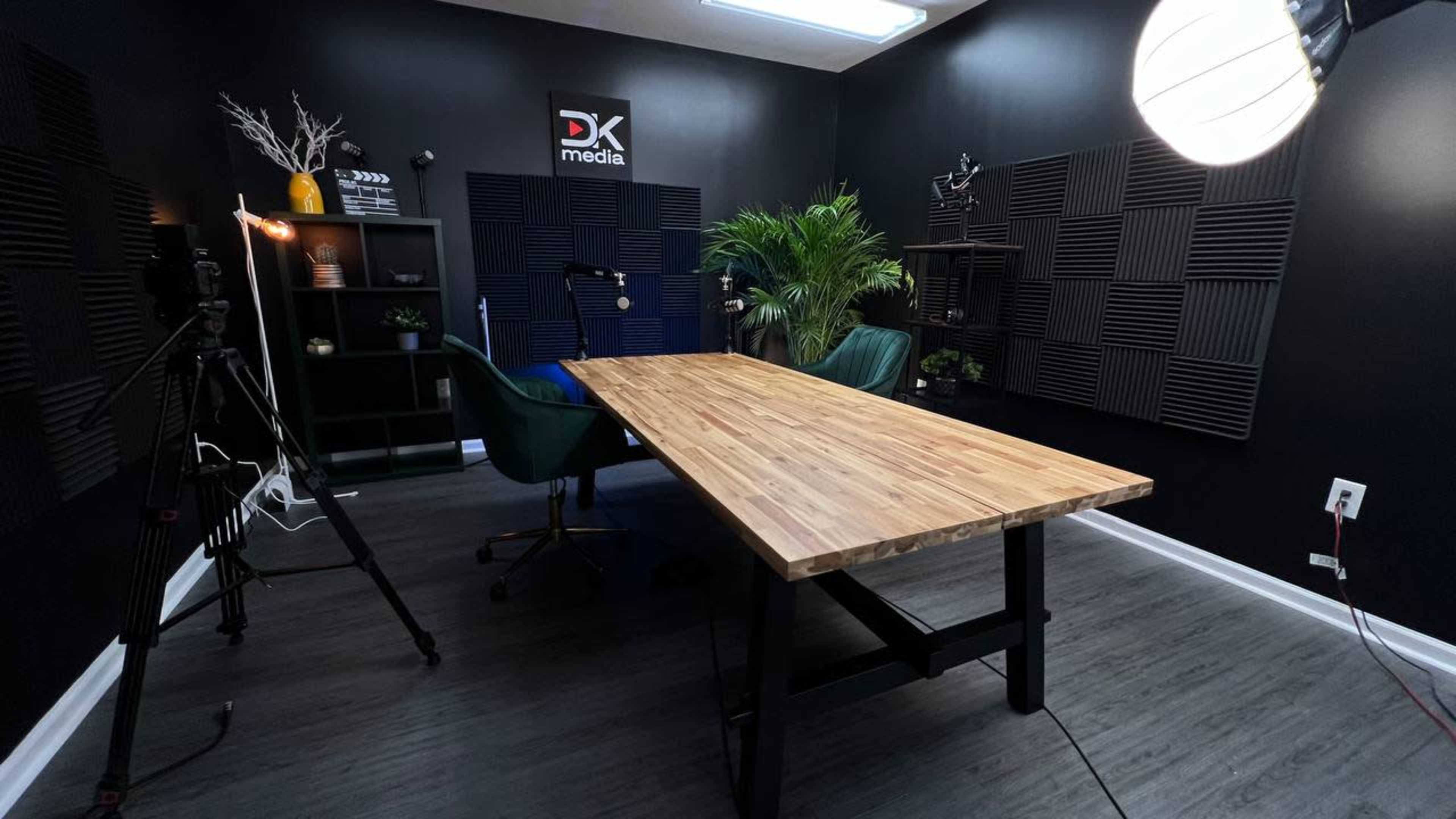 The image shows a well-lit recording studio with a large wooden table, two green chairs, and soundproofing panels on the walls.