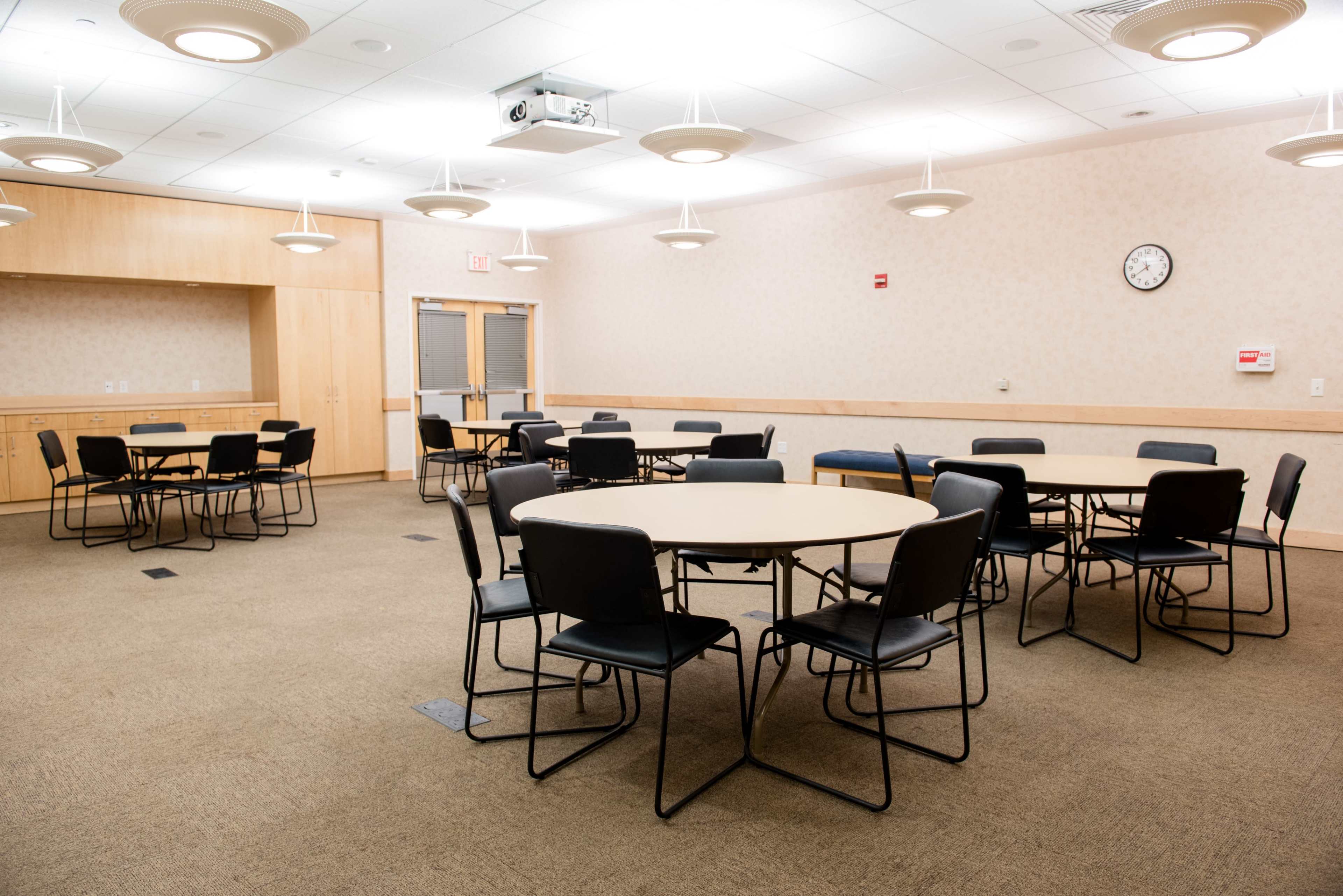 A spacious meeting room with several round tables and black chairs arranged on a beige carpet.