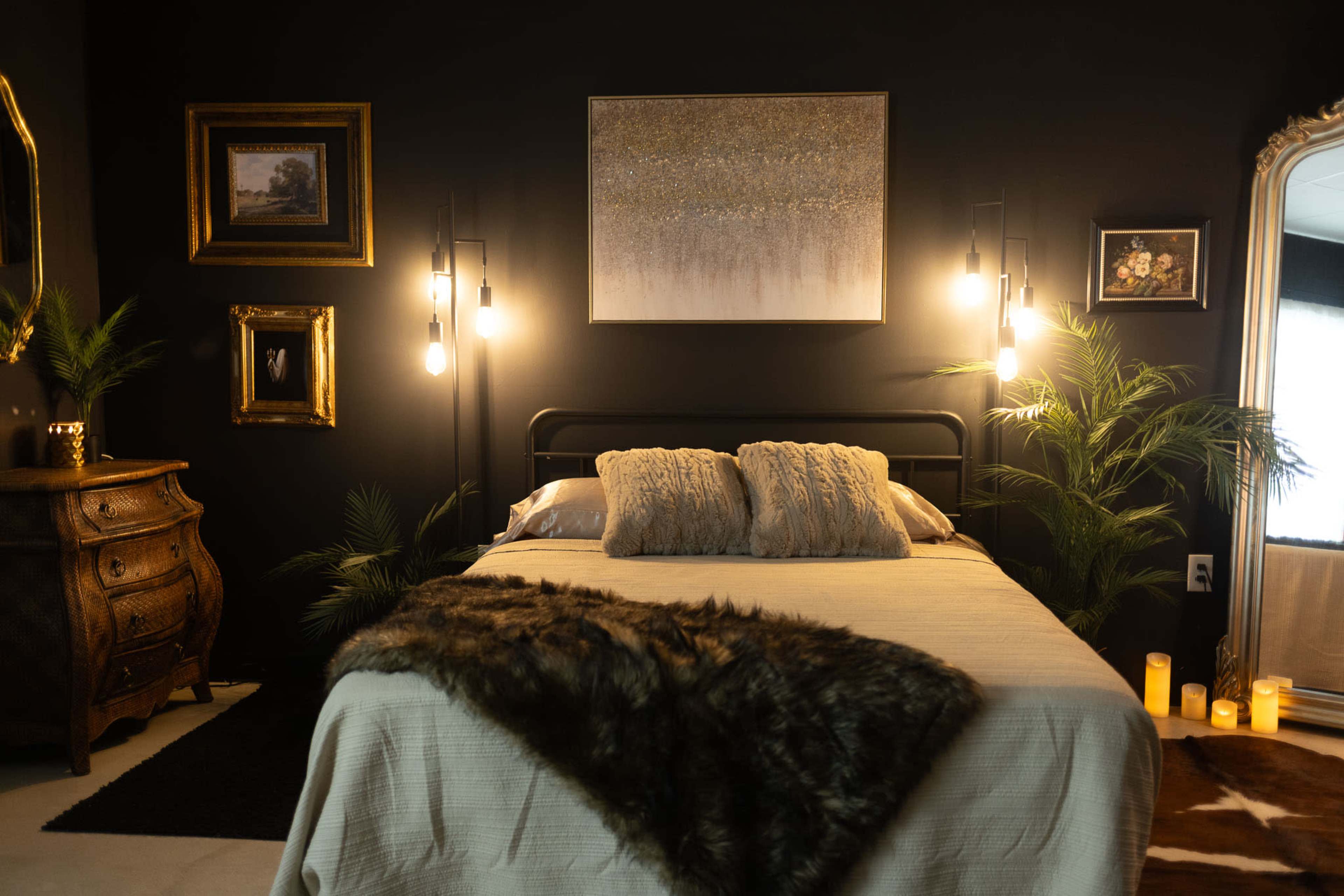 The image shows a darkly decorated bedroom featuring a bed with decorative pillows, framed artwork on the walls, and ambient lighting from hanging bulbs and candles.