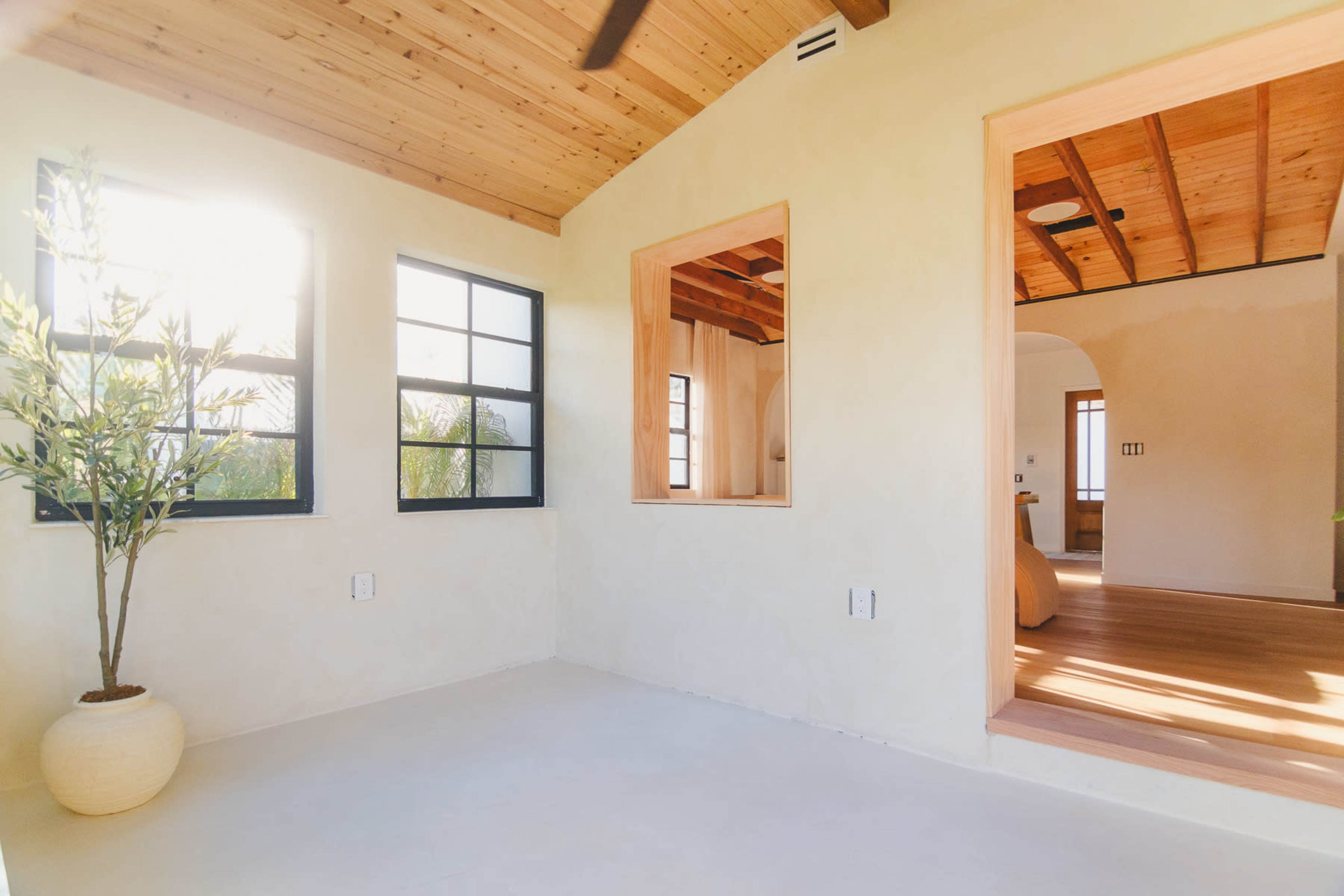 The image shows a bright and minimalist interior space featuring a plaster wall, large windows, and wooden ceilings.