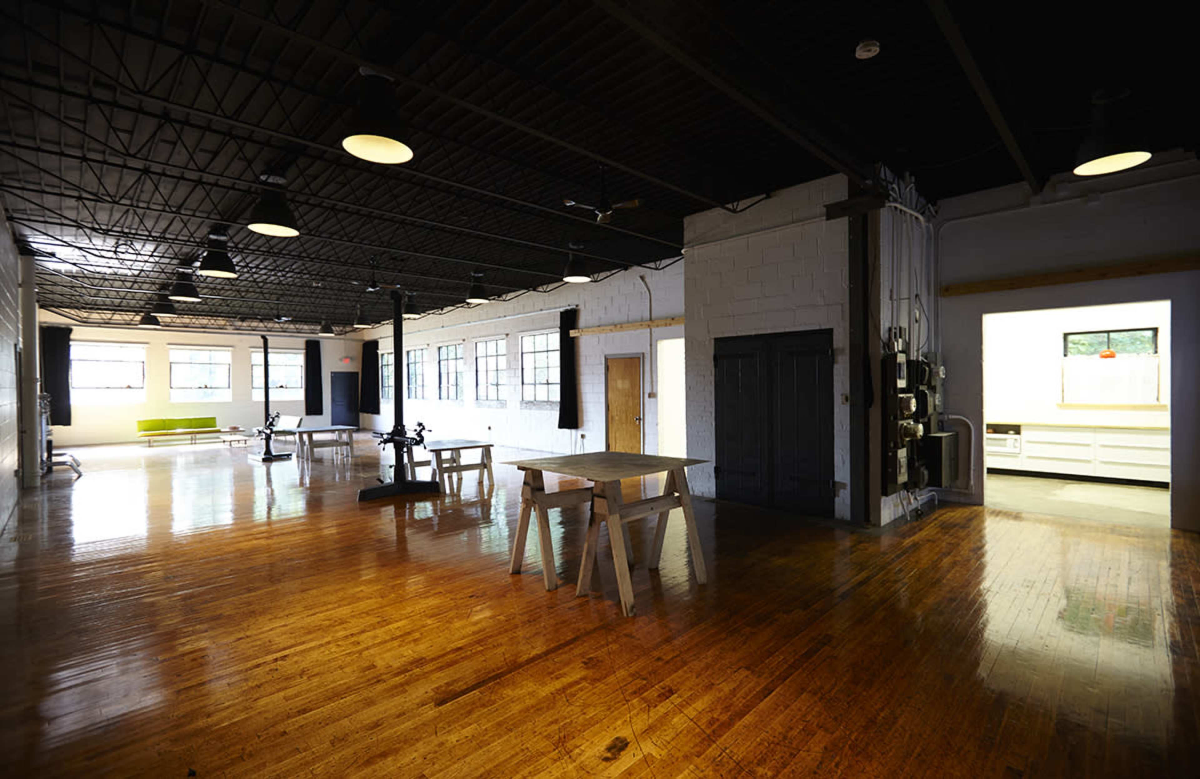 The image shows a spacious, empty industrial loft with polished wooden floors, large windows, and minimal furniture.