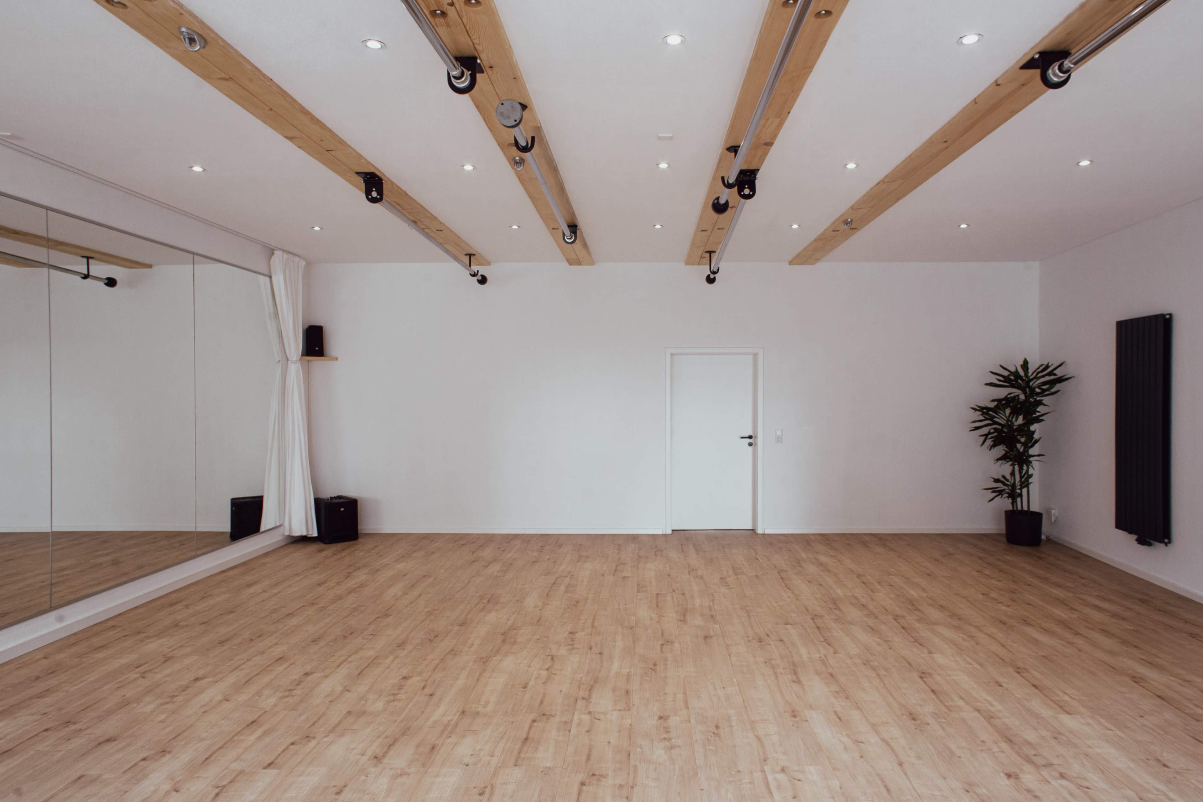 The image shows a spacious, empty room with wooden flooring, a large mirror on one wall, and a door leading to another area.