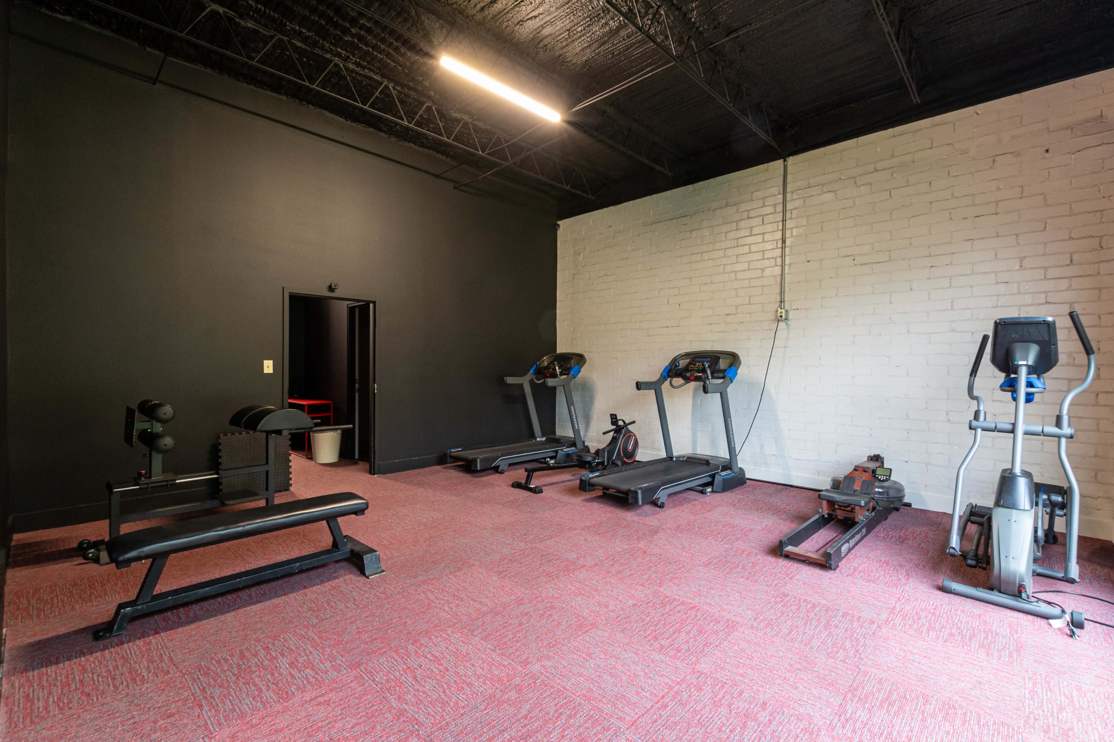 A fitness room with several exercise machines, including treadmills, a rowing machine, and a weight bench, on a carpeted floor.