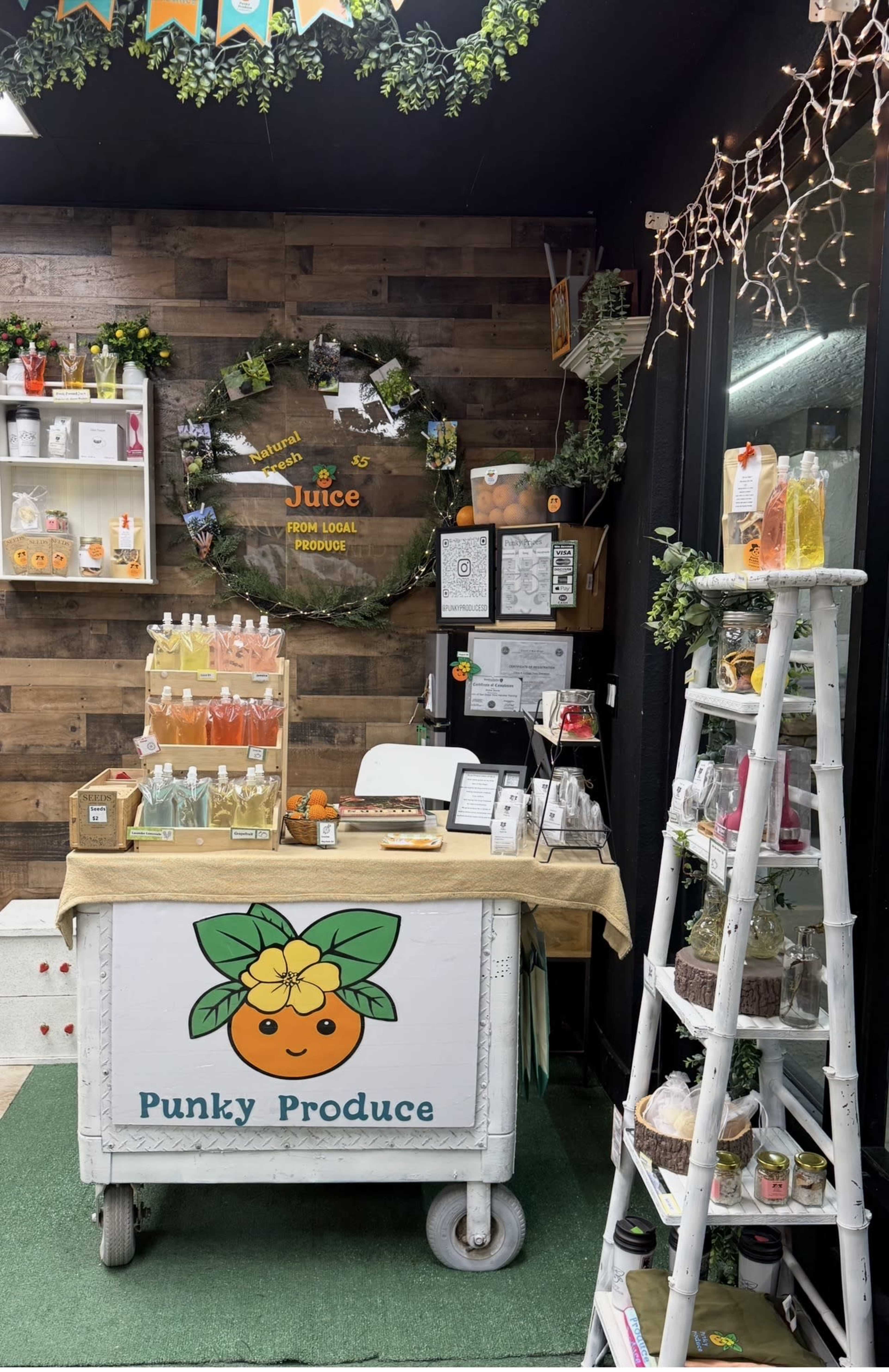 The image shows a small produce stand named "Punky Produce," featuring a variety of colorful juice bottles and produce items on display, set against a wooden backdrop with decorative lighting.