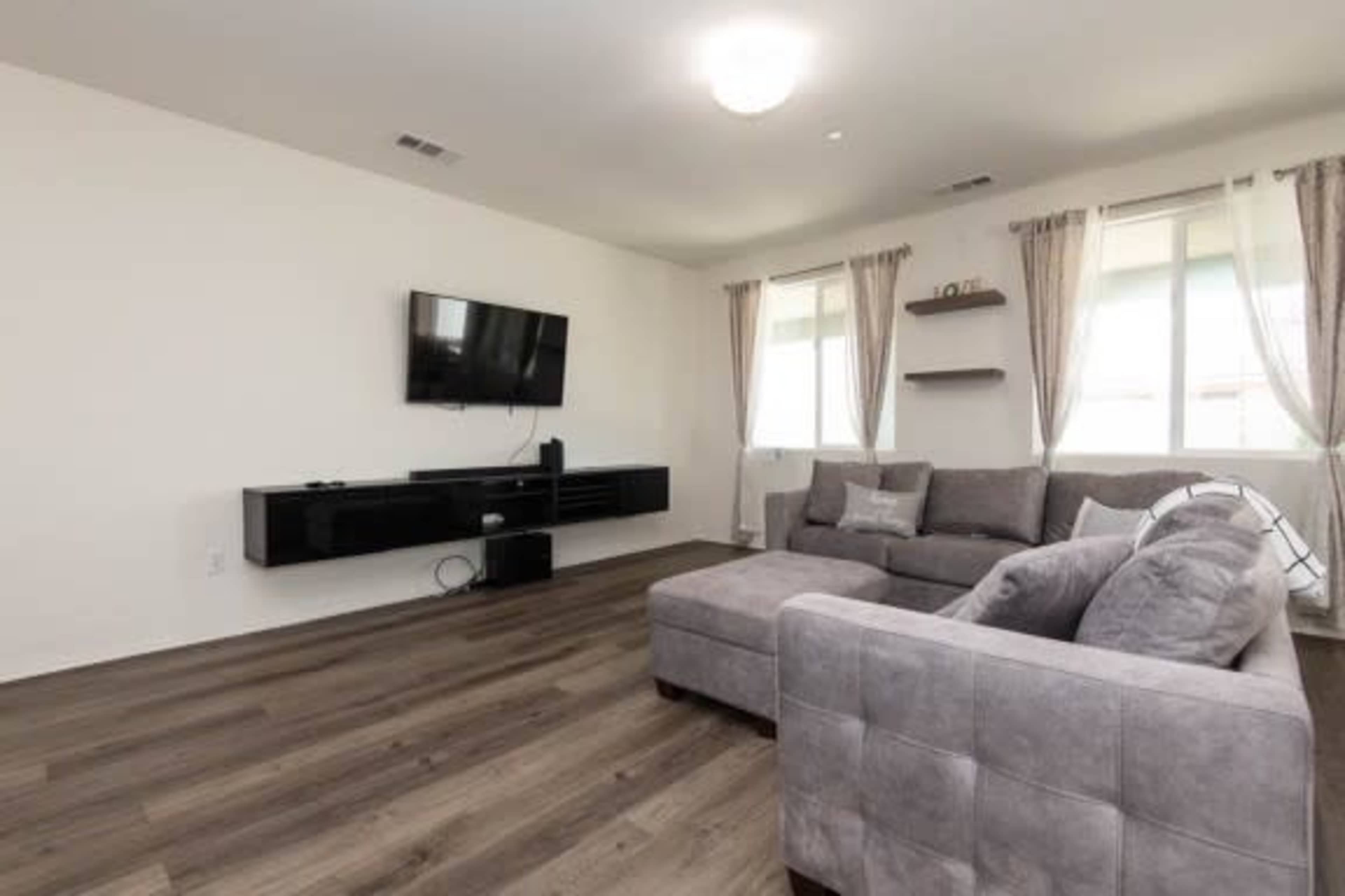 The image shows a modern living room with a gray sectional sofa, a wall-mounted television, and large windows dressed with light curtains.