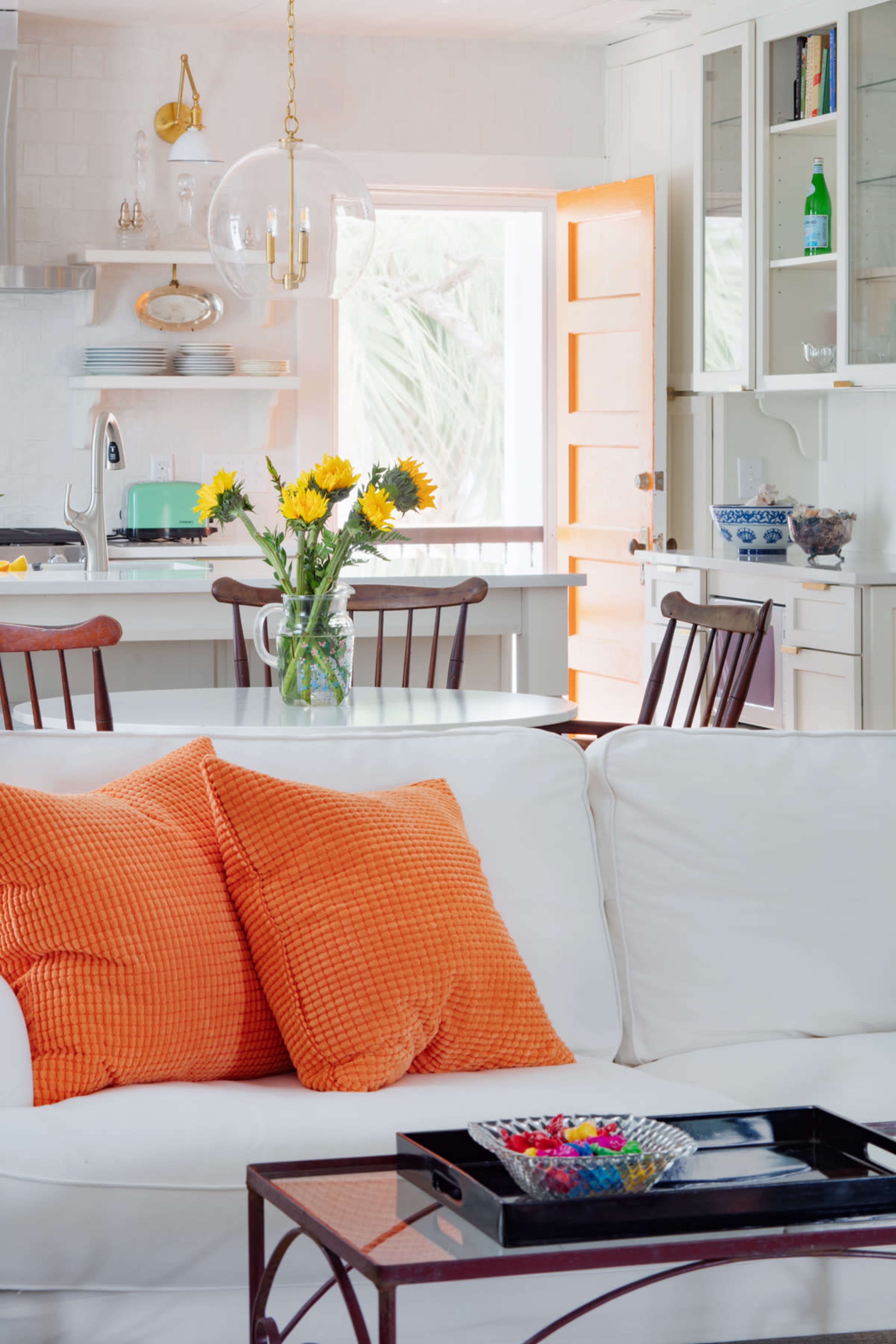 A white living room features a couch with orange pillows, a glass coffee table, and a kitchen area in the background with a window and a vase of sunflowers on the table.