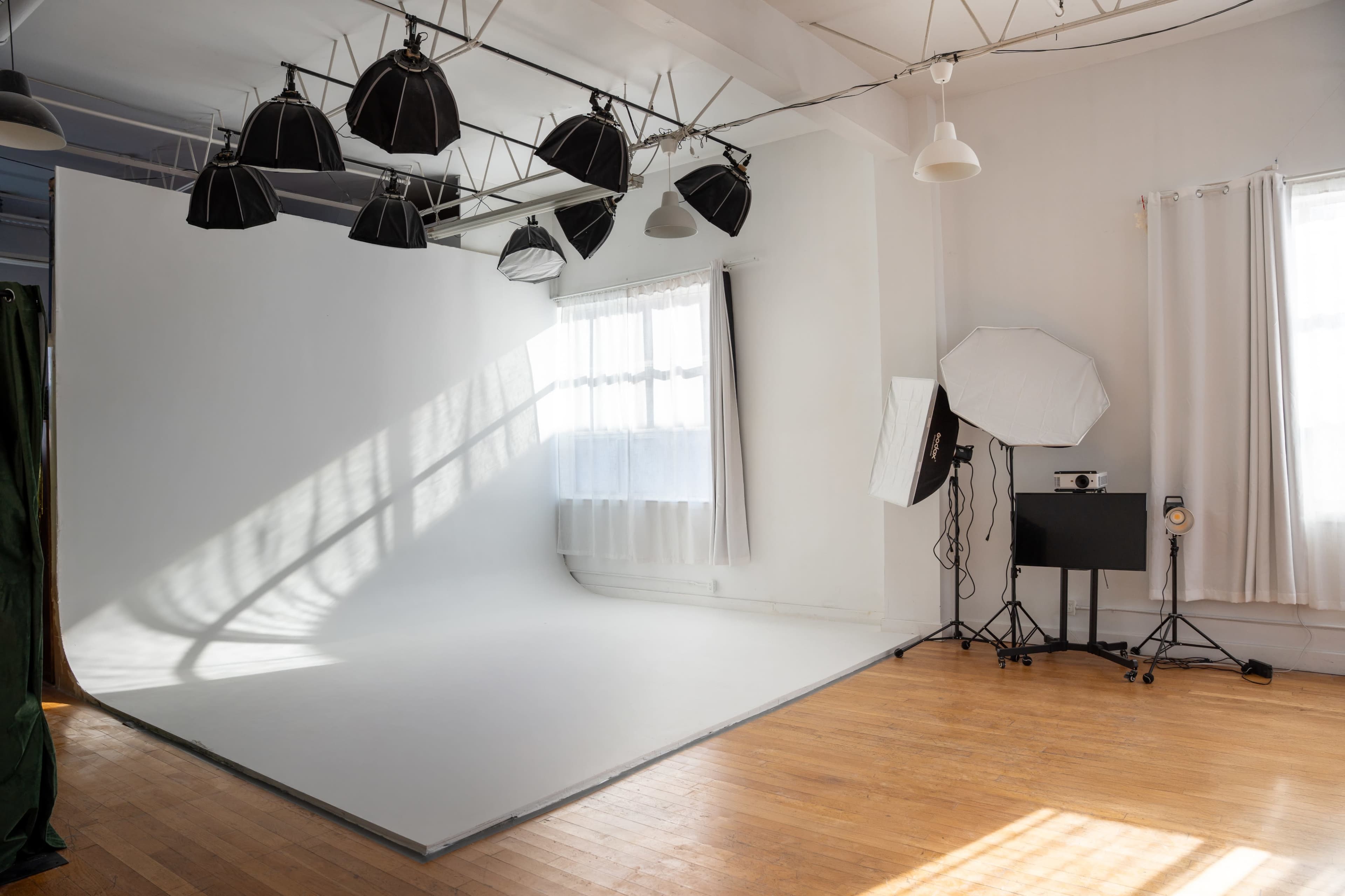 The image shows a well-lit photography studio with a large white backdrop, multiple softboxes hanging from the ceiling, and equipment set up in one corner.