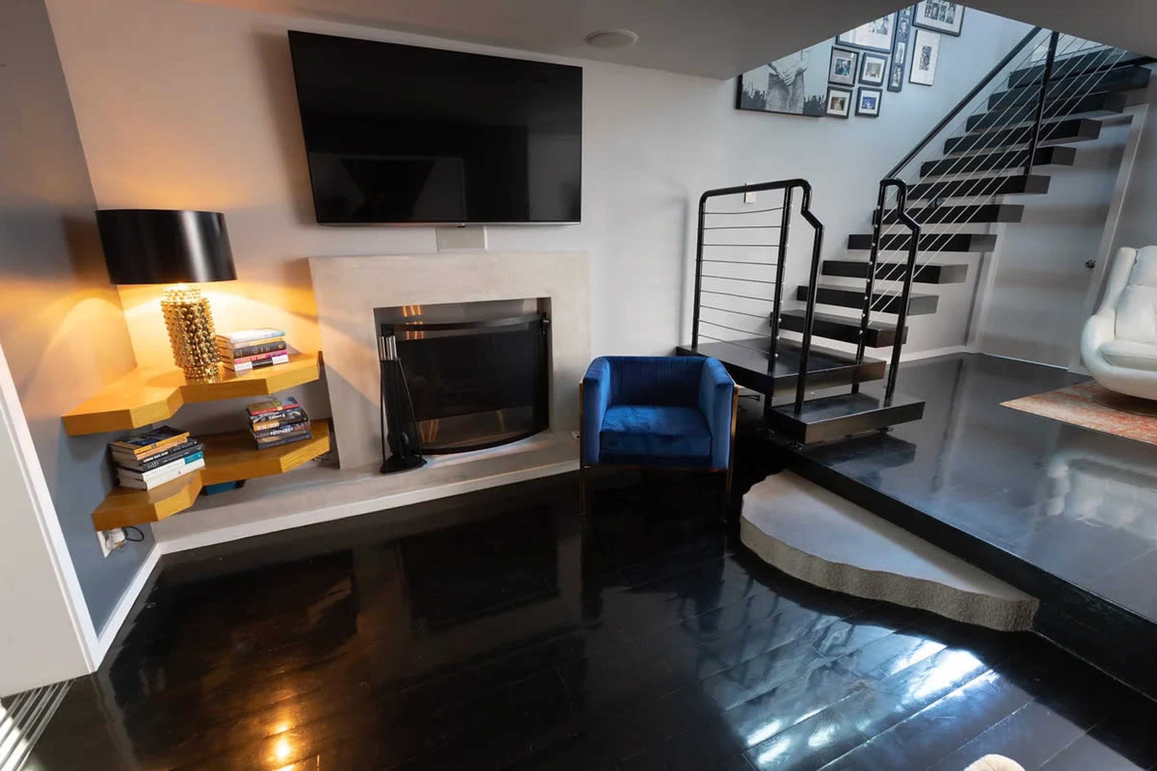 A modern living area with a flat-screen TV mounted above a fireplace, a blue chair, and a staircase leading to an upper level.