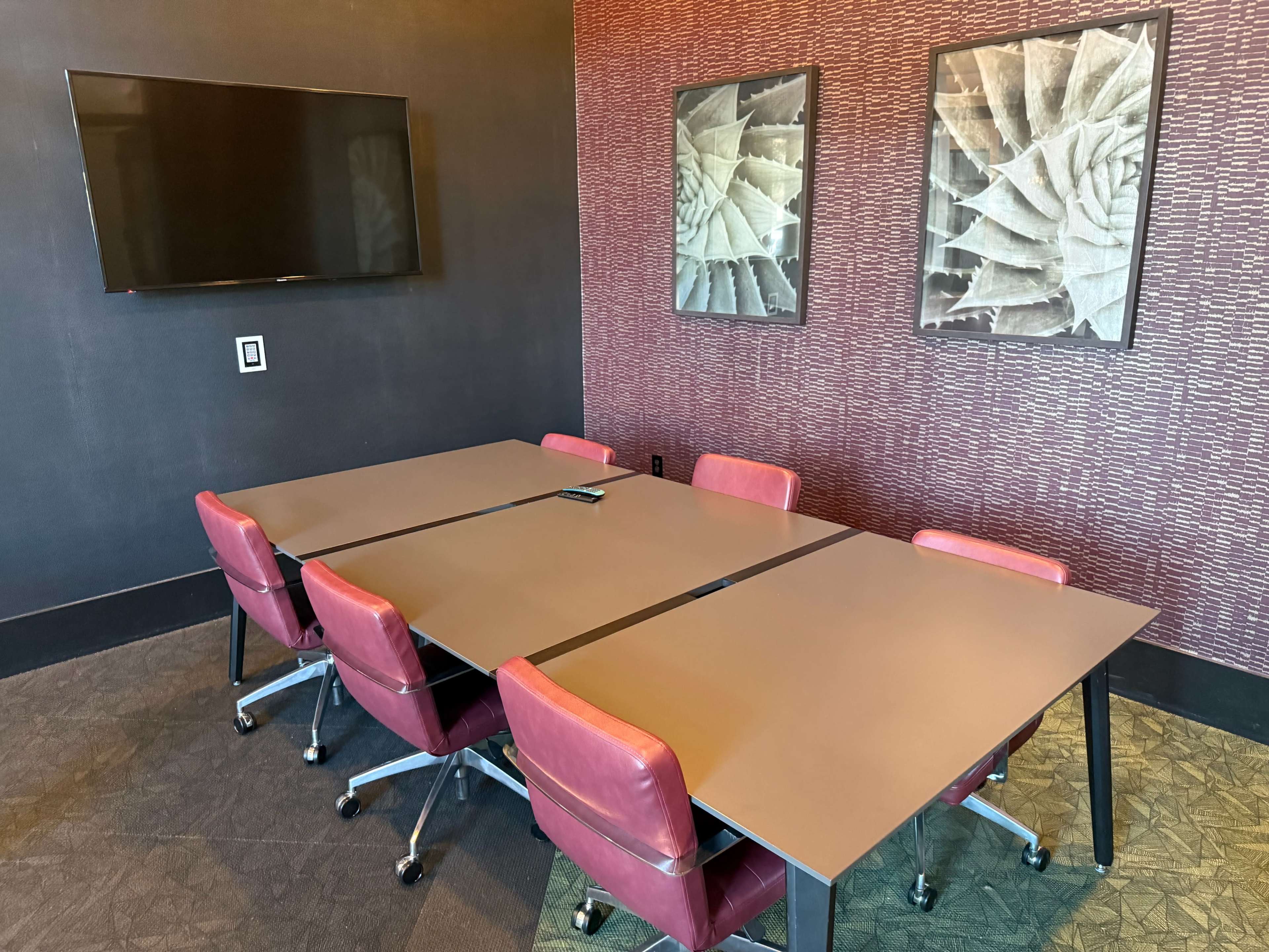A modern conference room features a rectangular table surrounded by red chairs, with a large wall-mounted screen and two framed prints on the walls.