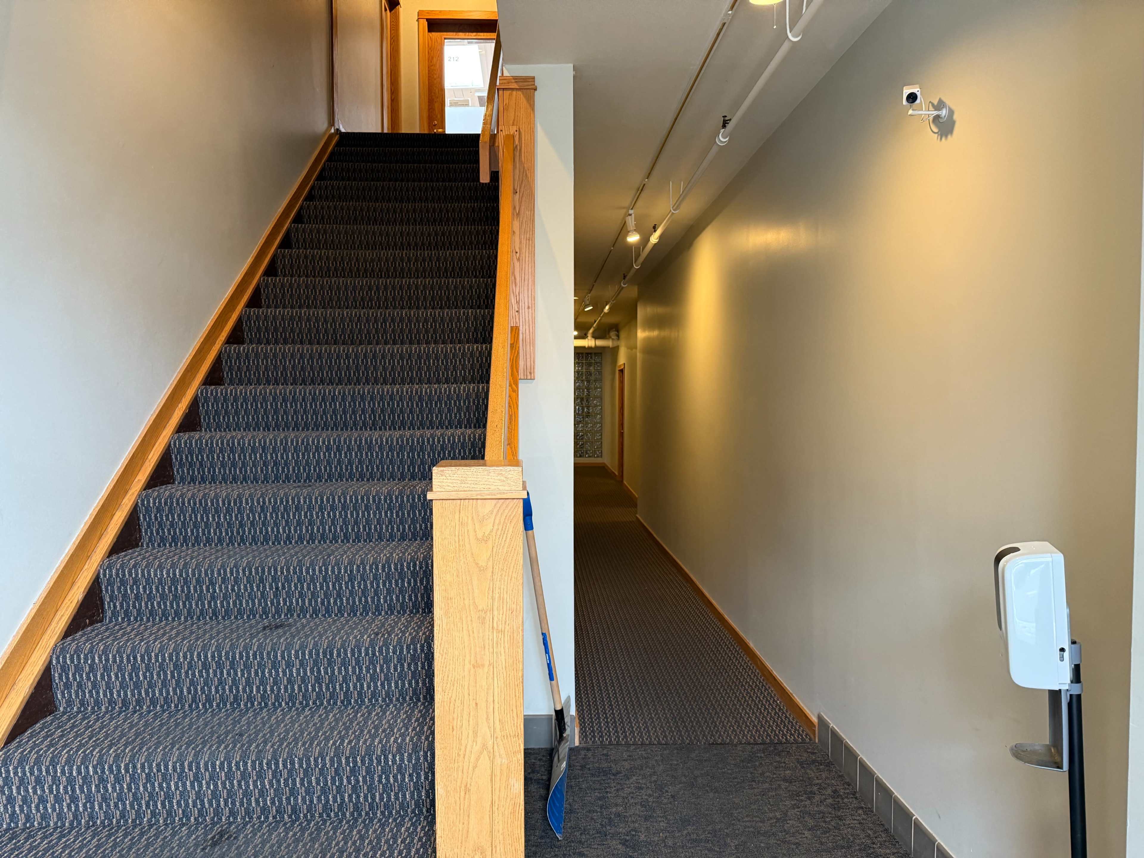 The image shows a carpeted staircase leading up on the left and a hallway with a wall-mounted camera and hand sanitizer dispenser on the right.