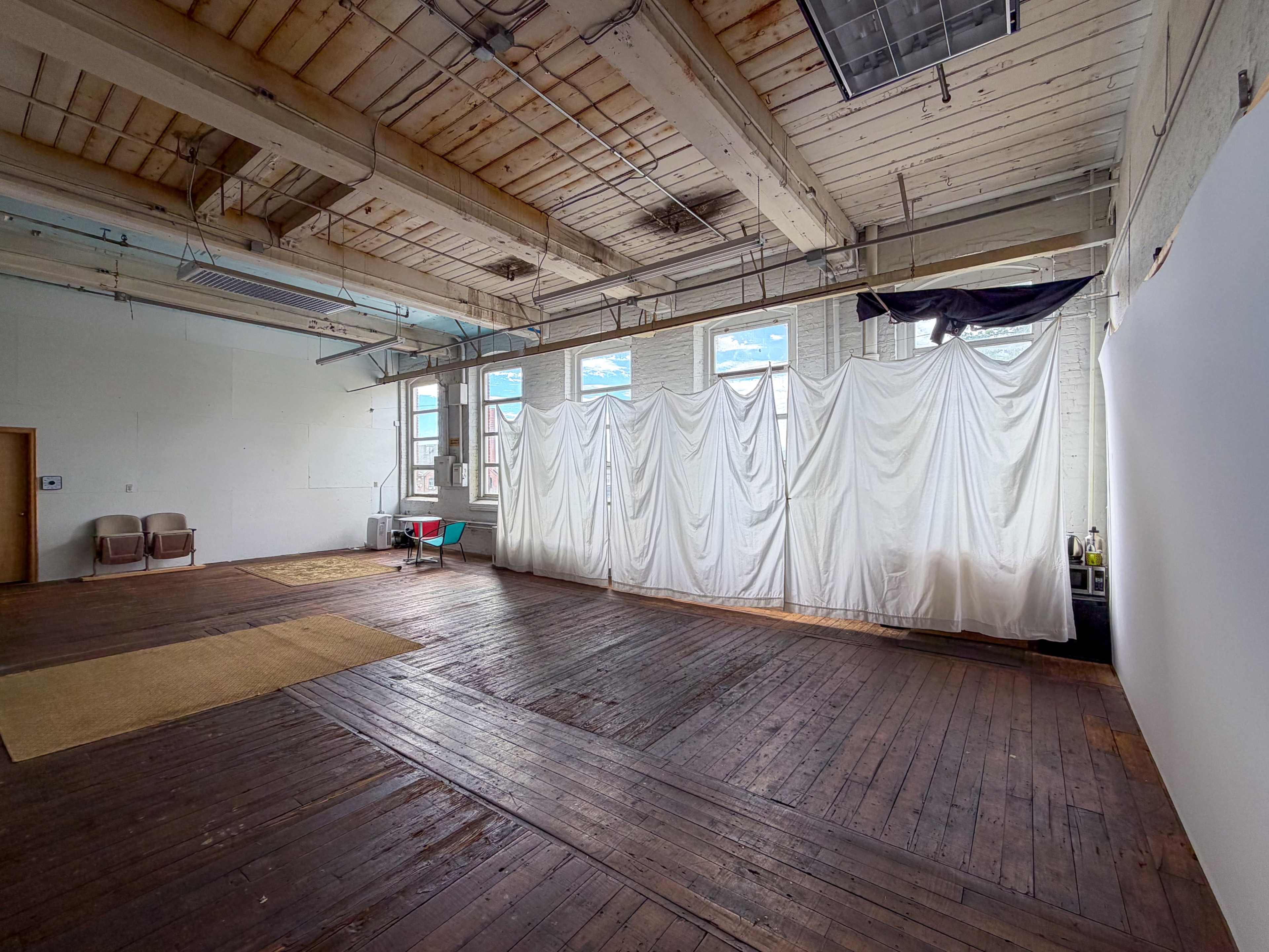 The image shows a spacious, empty room with wooden floors, large windows letting in natural light, and a white fabric draped in front of a wall.