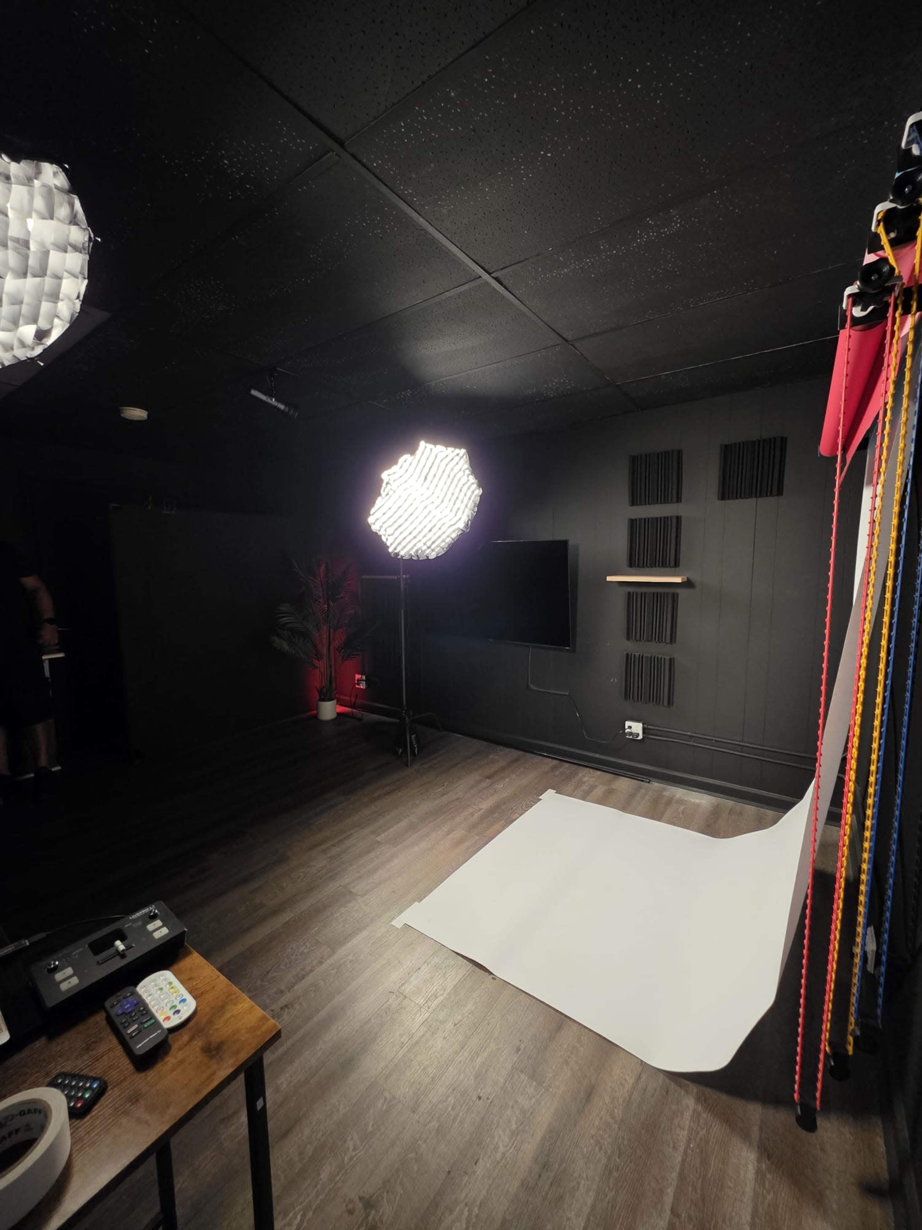 The image shows a photography studio with a white backdrop, lighting equipment, and a wooden table with various tools and remotes.