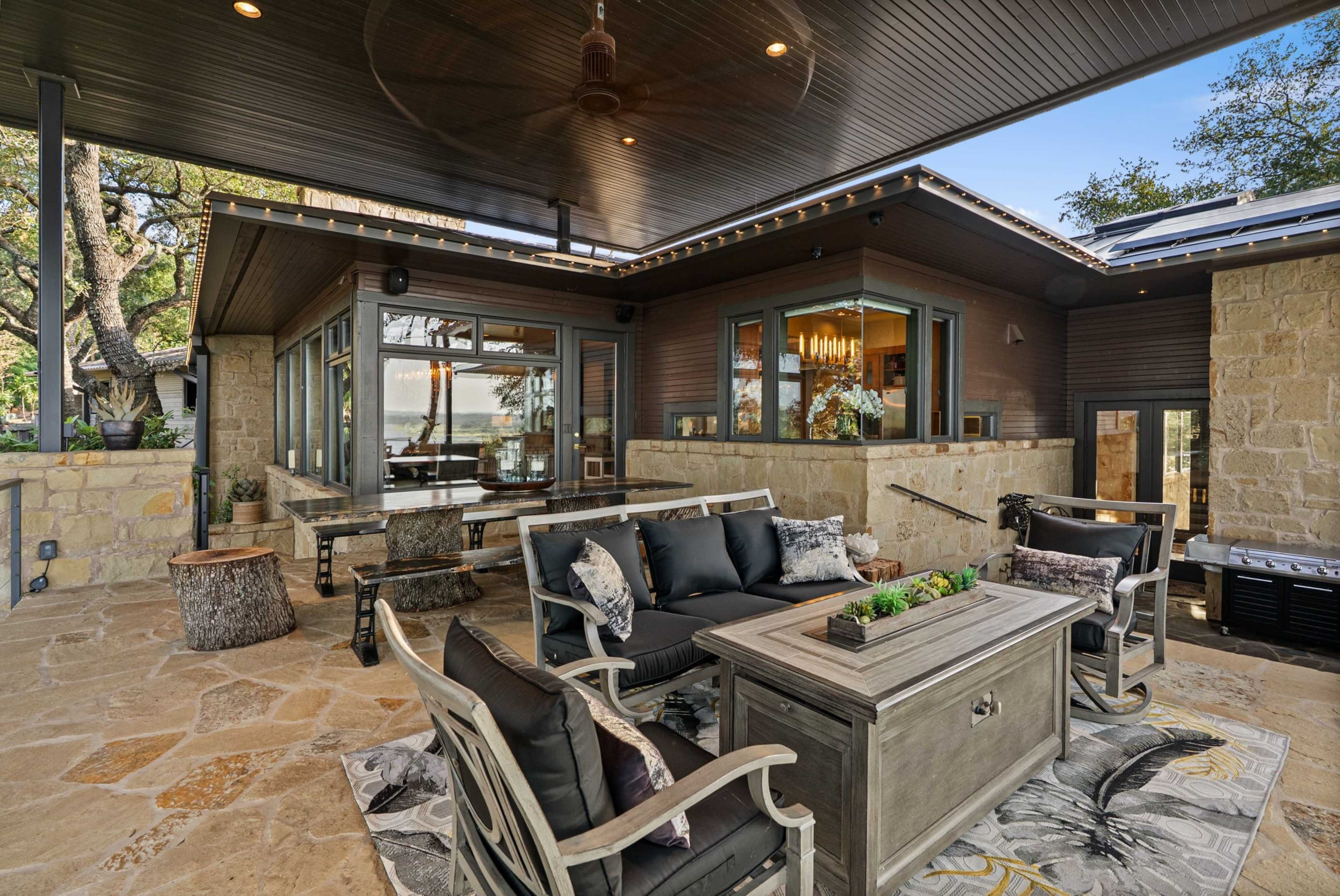 The image shows a spacious outdoor living area with stone flooring, comfortable seating, and large windows overlooking a scenic view.