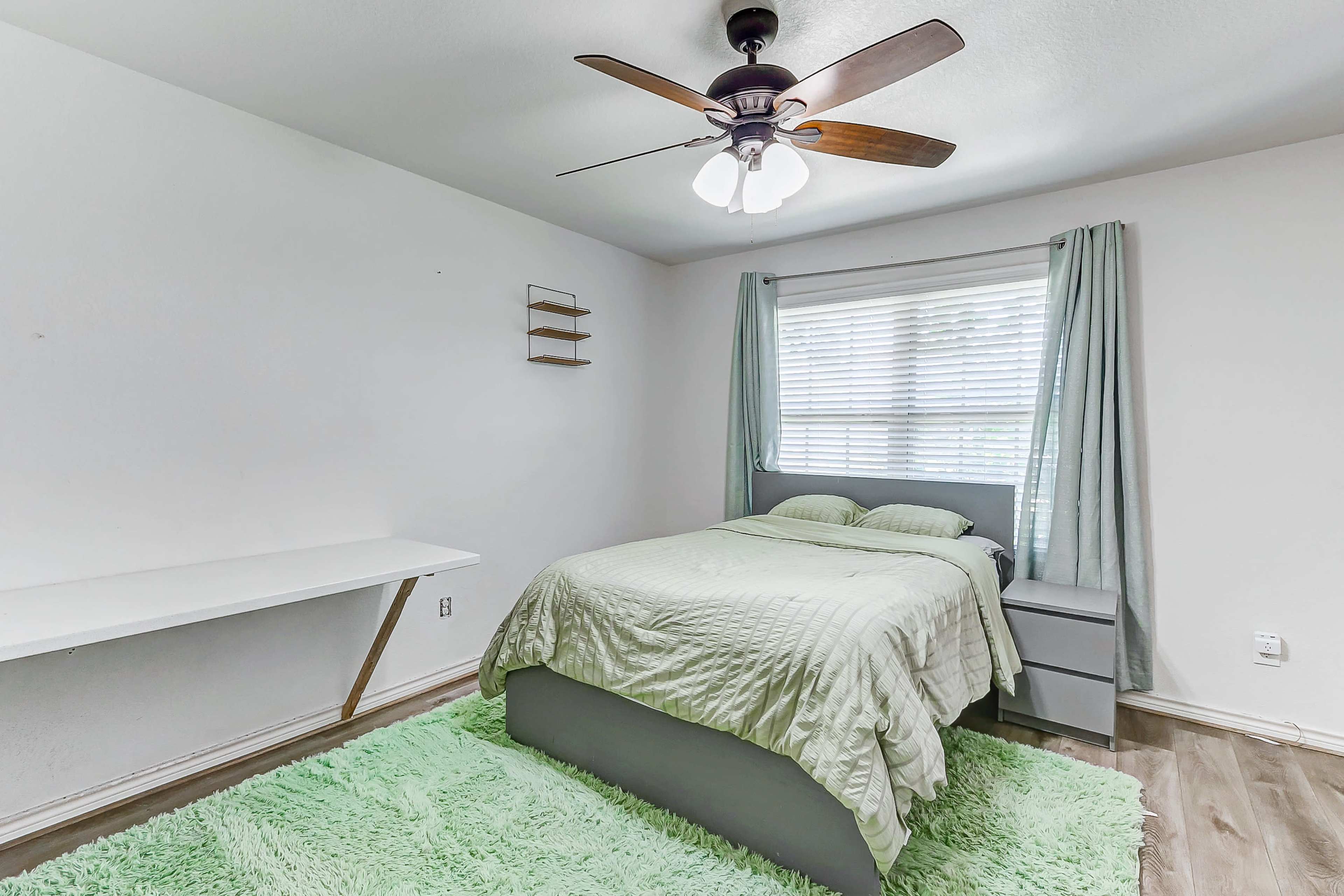 A cozy bedroom featuring a bed with light green bedding, a wooden ceiling fan, a window with sheer curtains, and a desk next to a fluffy green rug.