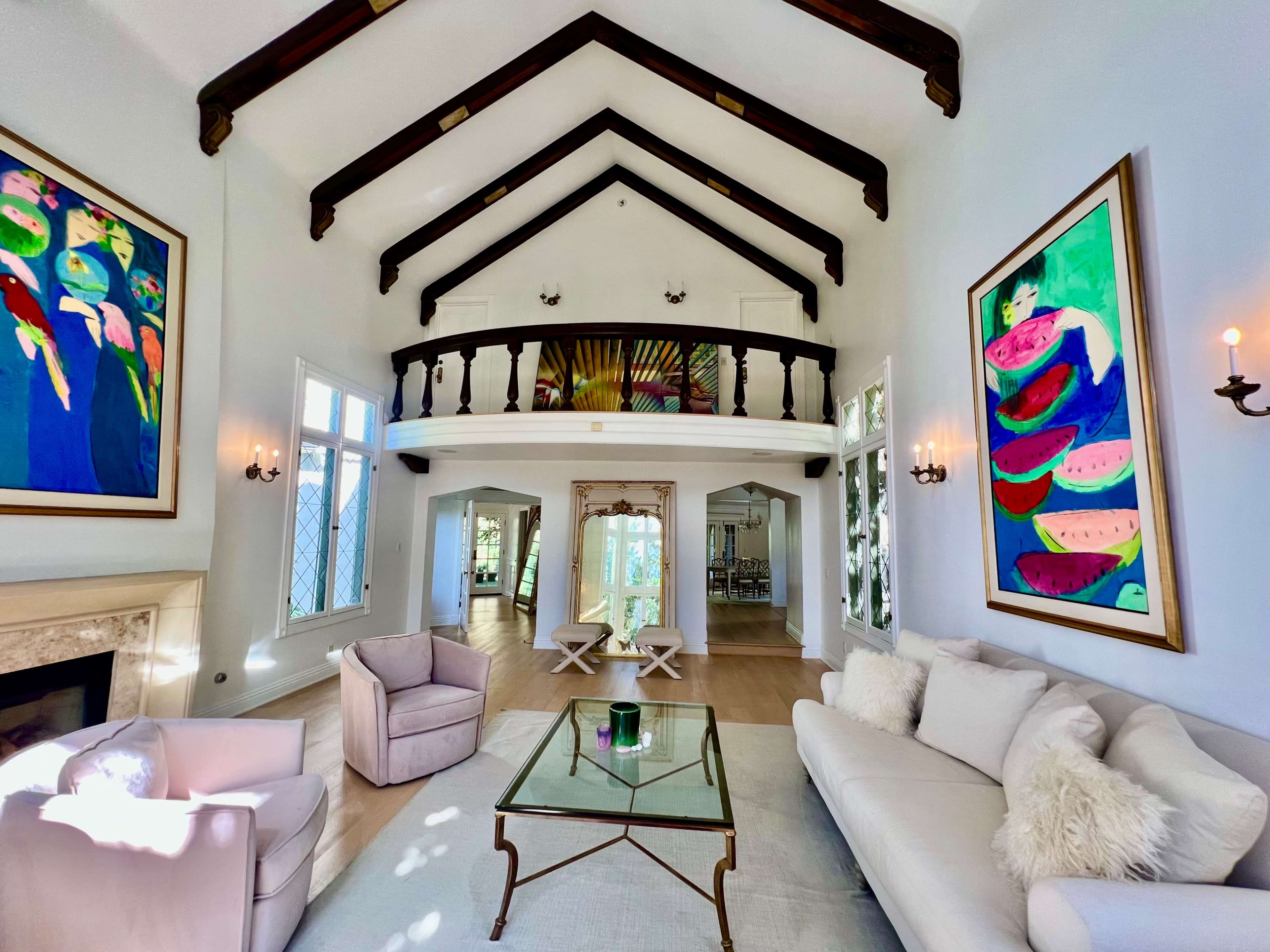 A bright living room with high vaulted ceilings, large windows, and a comfortable seating area featuring a sofa and armchairs, complemented by colorful artwork on the walls.
