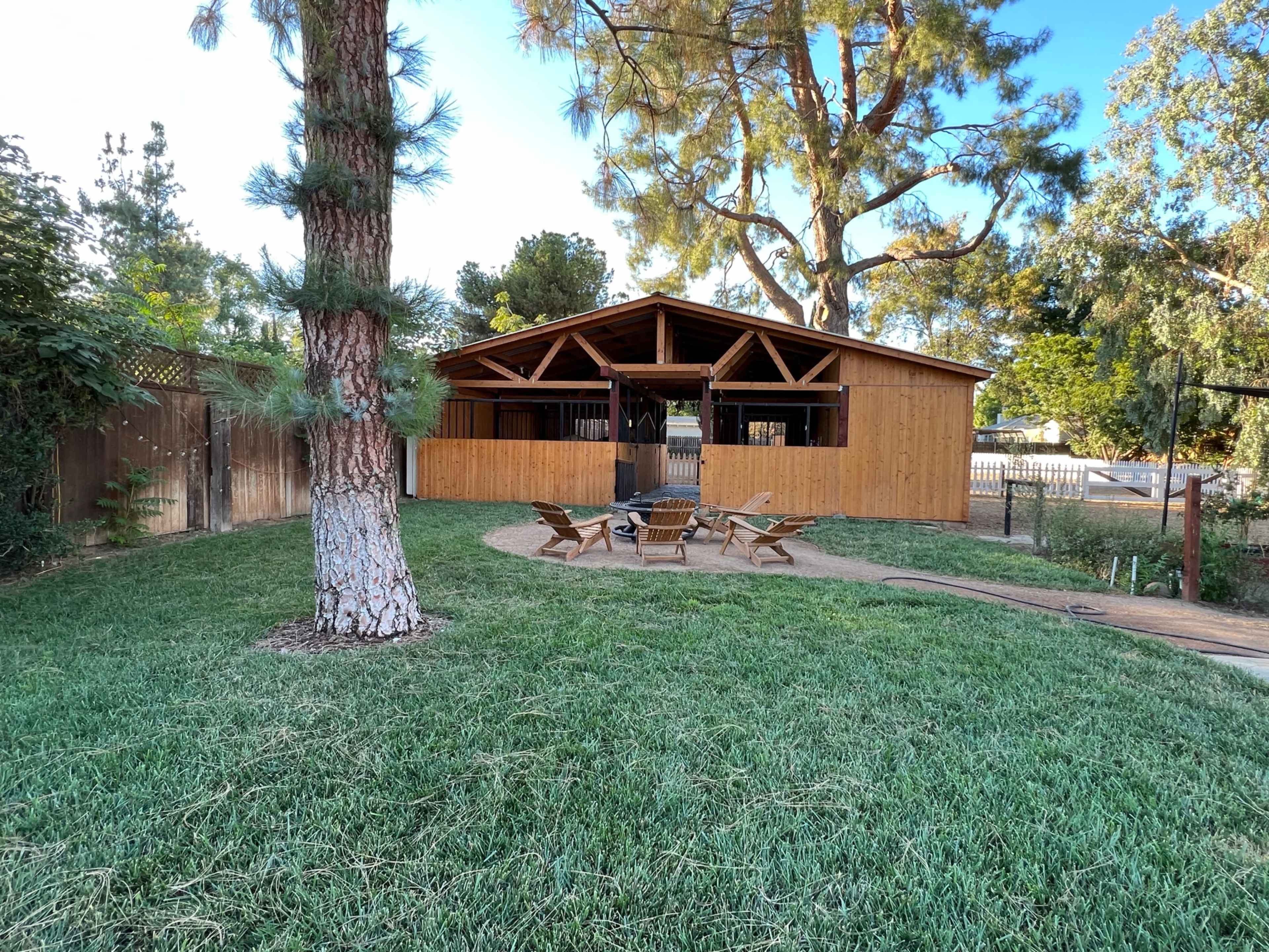 A wooden structure with a pitched roof sits in a grassy yard, surrounded by trees and a fire pit area with chairs.