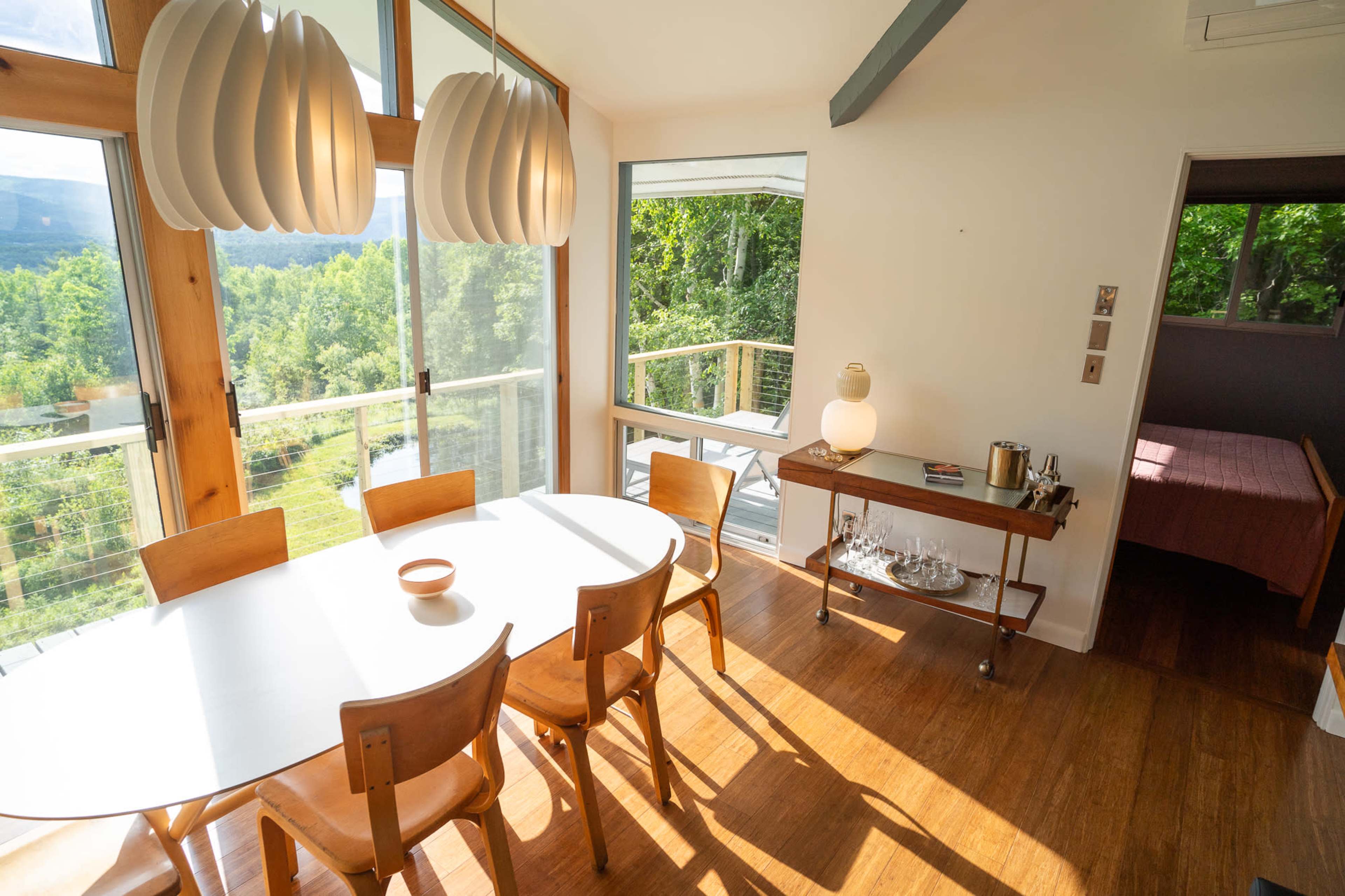 The image shows a modern dining area with a wooden table and chairs, large windows overlooking a green landscape, and a small bar cart nearby.